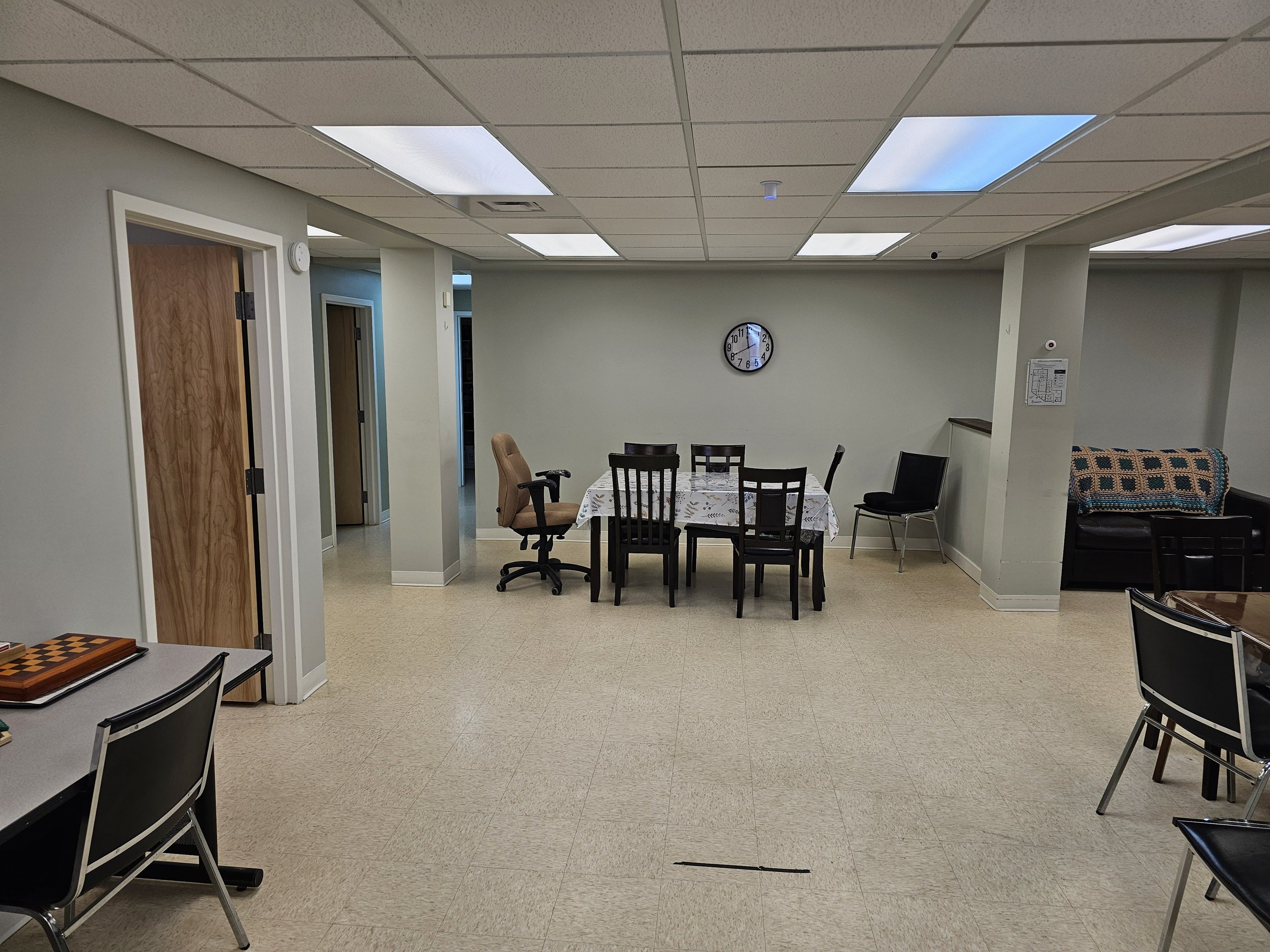 An empty multi-purpose room with beige tiled floor, white walls, a ceiling with fluorescent lights and skylights. In the center, a table with a white patterned tablecloth and five chairs. To the right, a small sitting area with a black sofa, an armchair, and more chairs. On the wall behind the table, a round wall clock shows approximately 12:06.