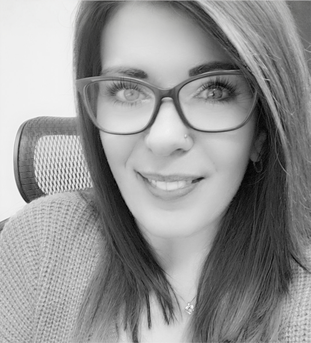 A black-and-white photo of a smiling woman with long hair, wearing glasses and a nose piercing, seated in front of a textured chair.