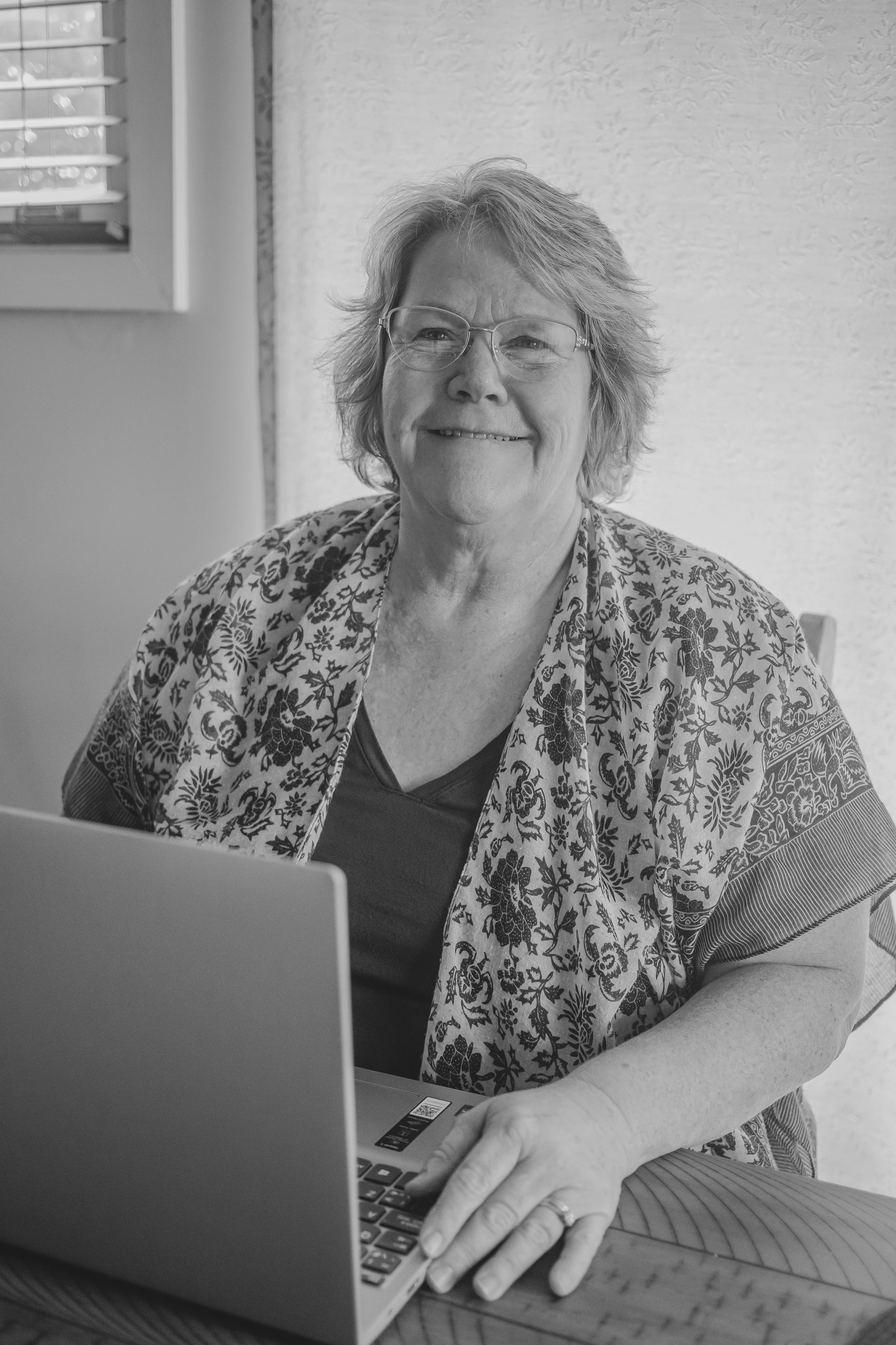 A smiling elderly woman with glasses sitting at a table with a laptop, wearing a patterned shawl over a dark shirt, in a room with a window and textured wall.