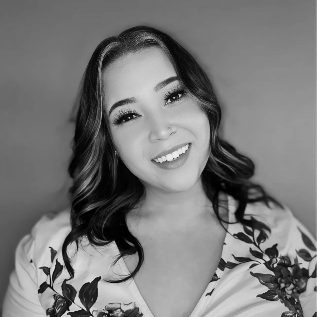 Black and white portrait of a smiling woman with wavy dark hair and makeup, wearing a floral-patterned top.