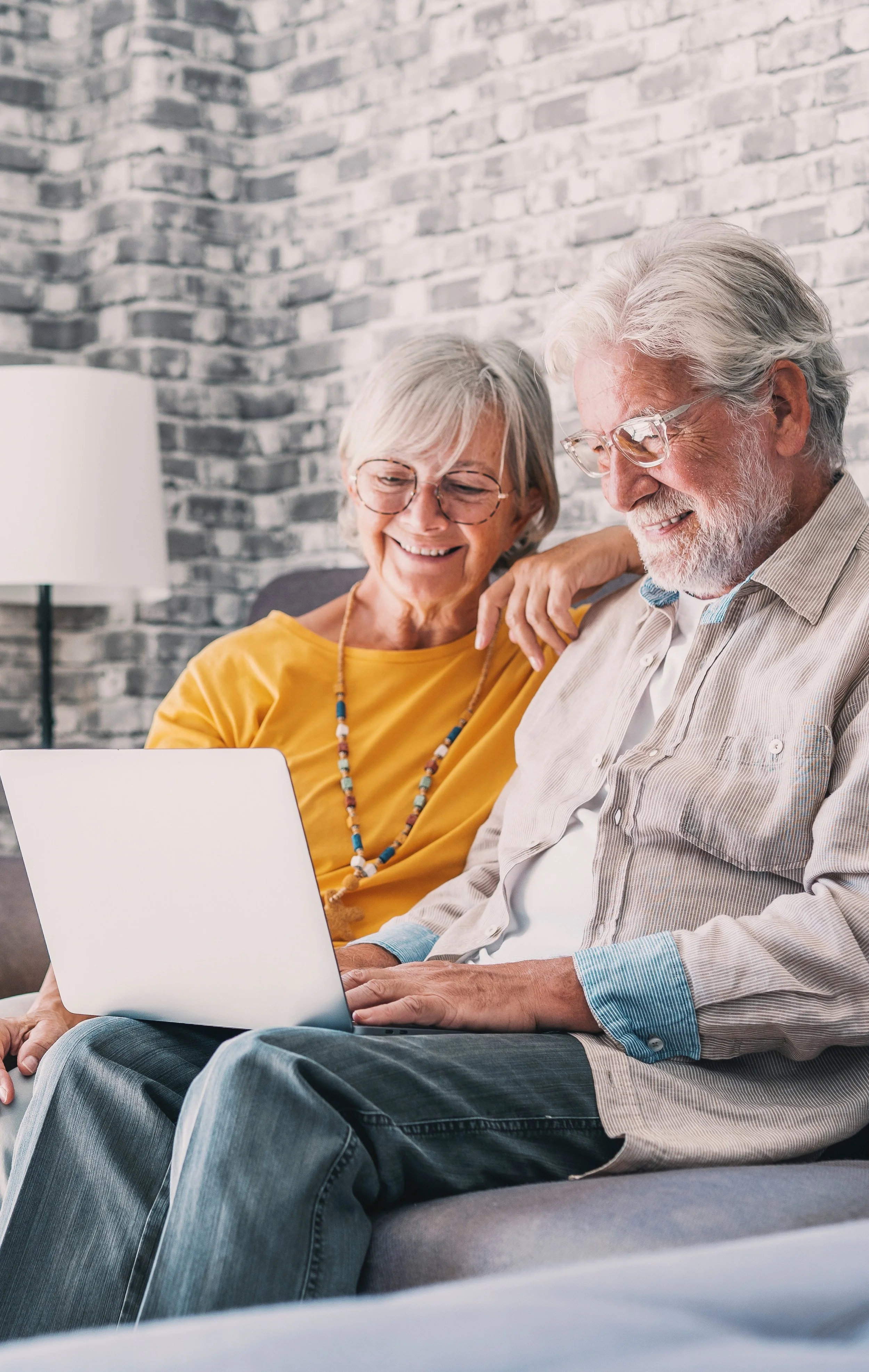 An elderly couple sitting on a gray sofa, looking at a laptop with smiles.