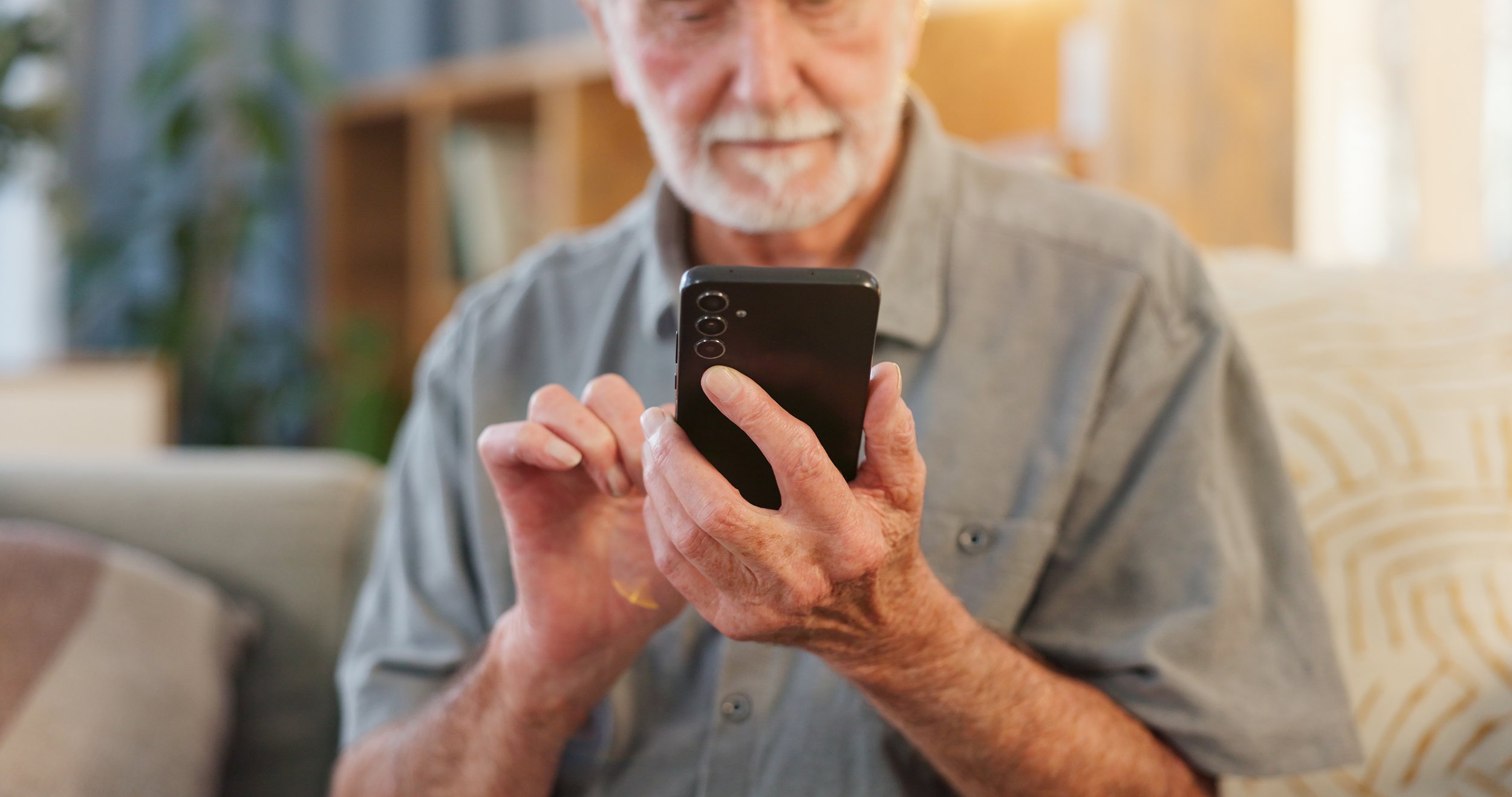 An elderly man with a beard is smiling and looking at his smartphone, sitting on a sofa in a well-lit room.
