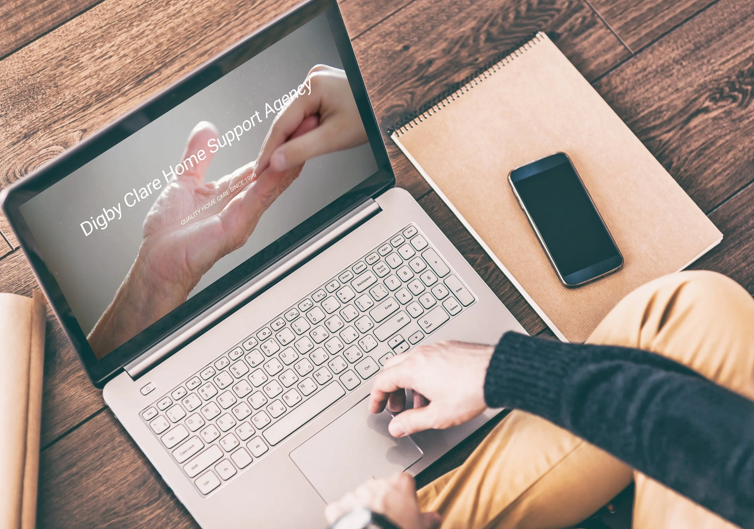 A person using a laptop with the screen displaying 'Digby Clare Home Support Agency'. The person is sitting at a wooden table with a smartphone and a notebook nearby.
