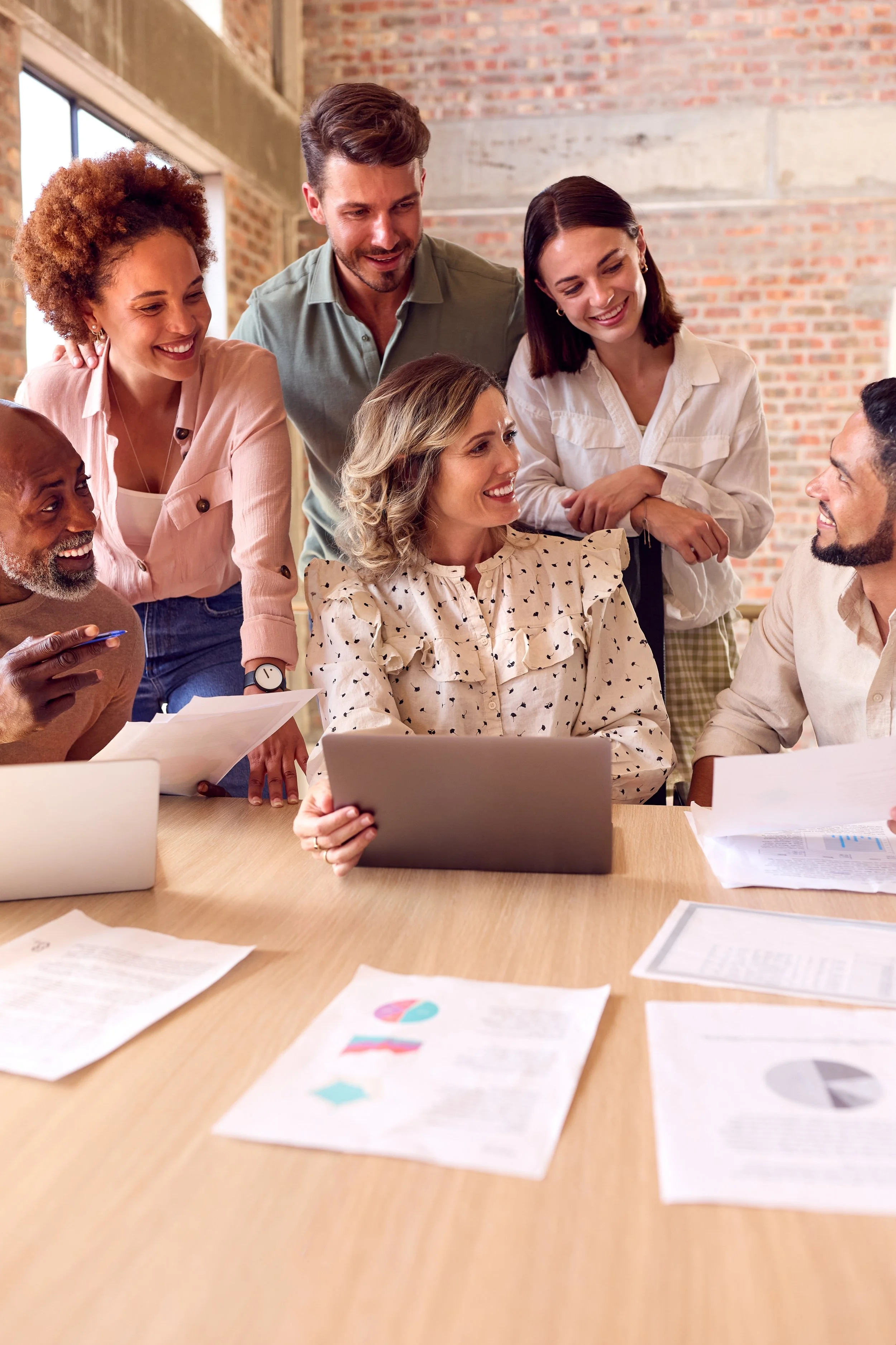 Group of six diverse professionals gathered around a table in a modern office, discussing documents and working on laptops, smiling and engaging with each other.