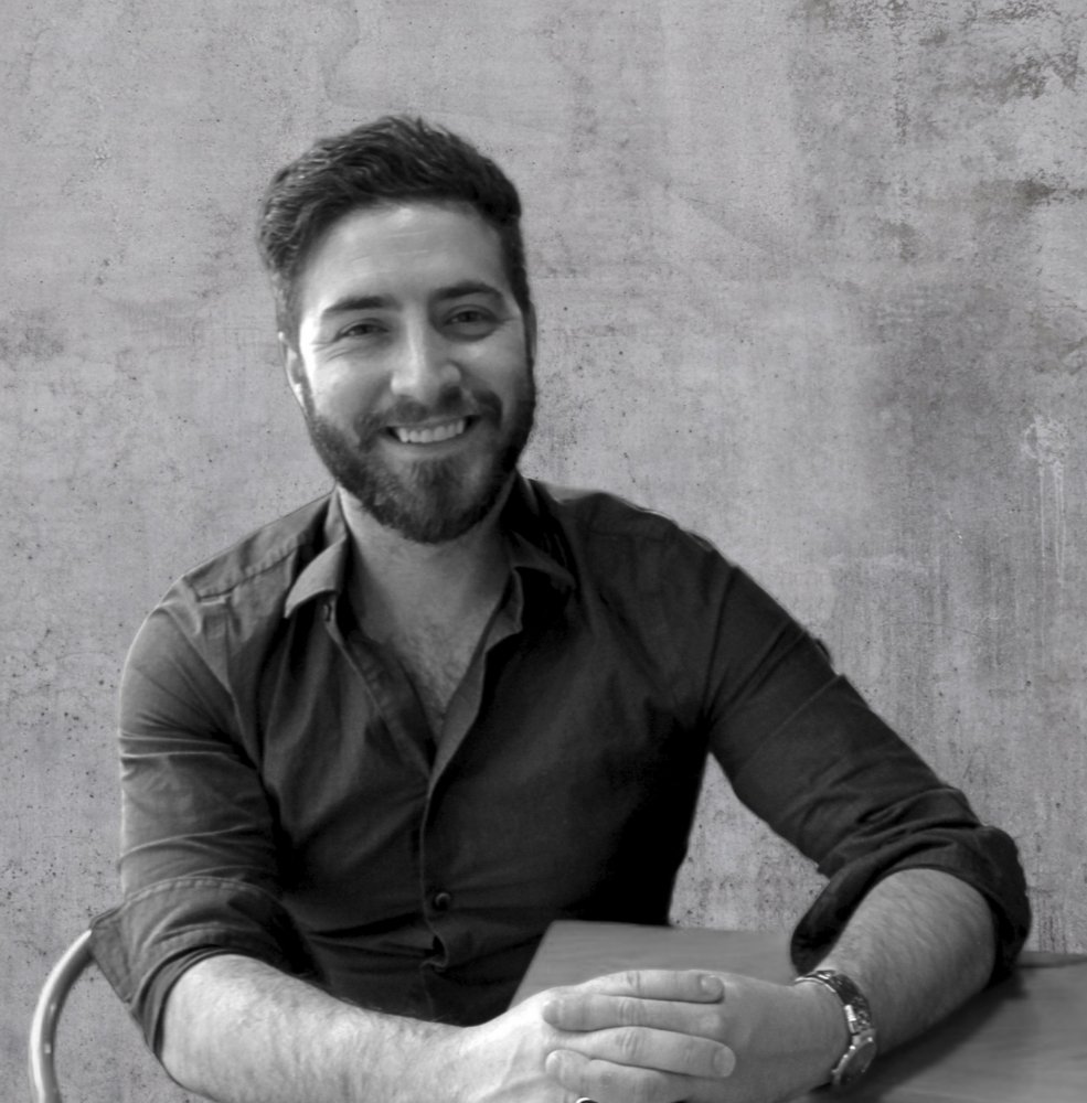 Black and white photo of a man sitting at a table, smiling, with a plain textured wall in the background.