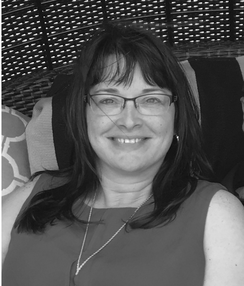A woman with glasses and shoulder-length hair smiling, sitting on a woven outdoor chair with decorative pillows.