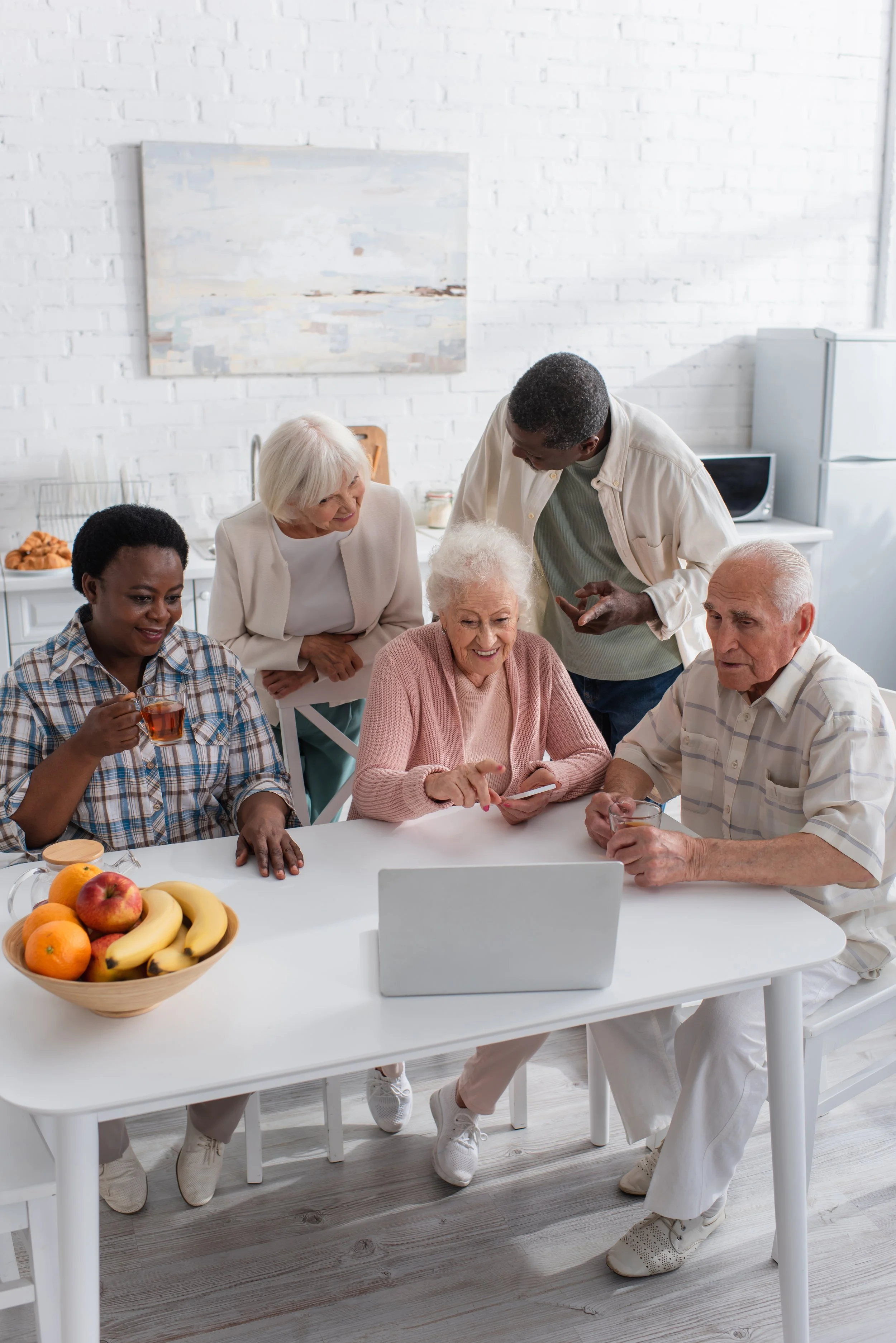 Group of five diverse elderly people gathered around a table, looking at a laptop screen and sharing a moment of laughter in a bright, modern kitchen.