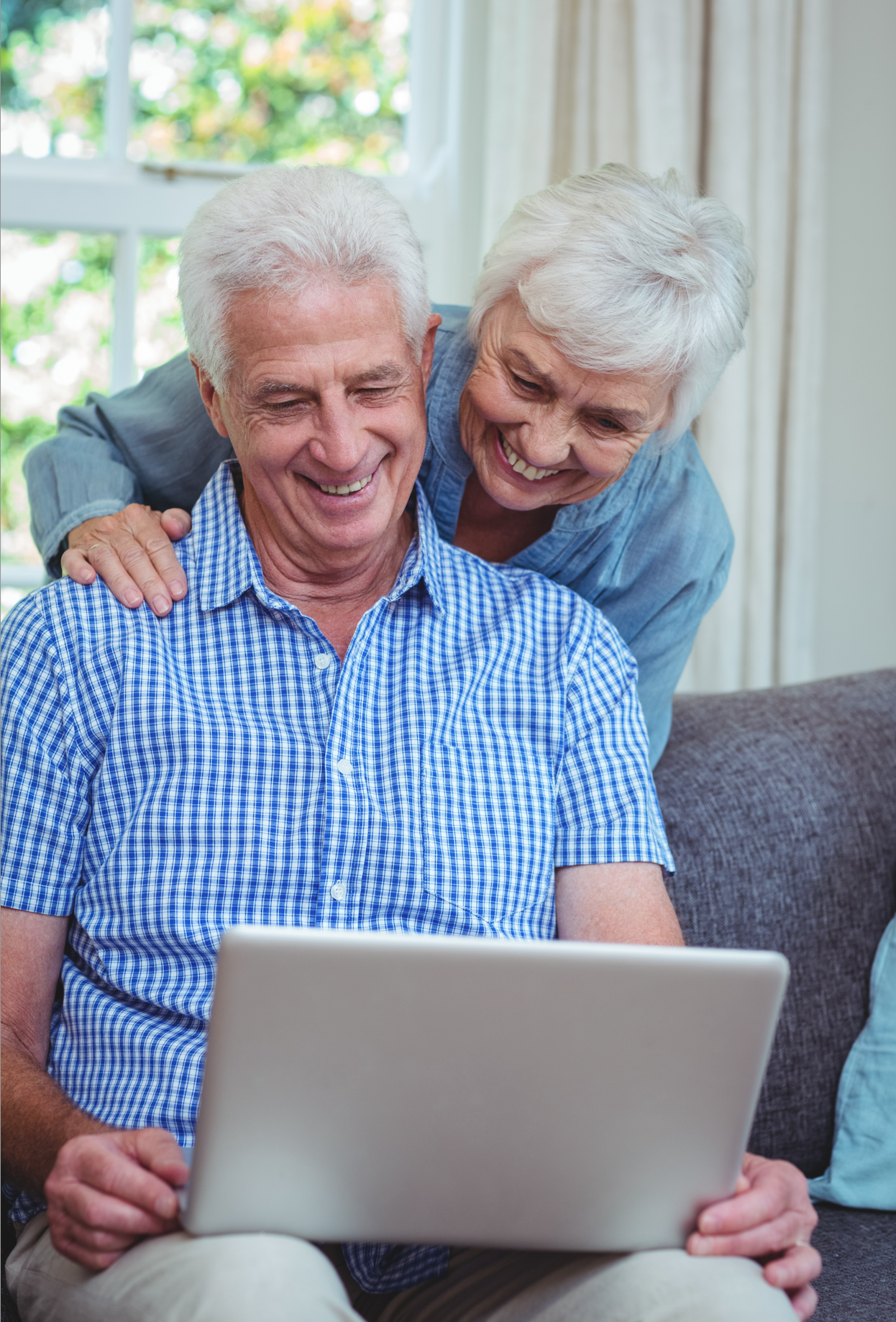 An elderly man sitting on a couch using a laptop, smiling, with an elderly woman leaning over and smiling behind him, in a well-lit room with a window and curtains.