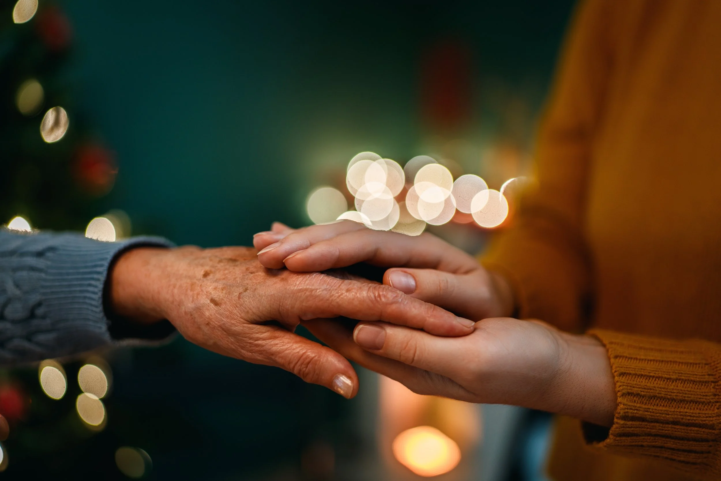 Two people, one elderly and one young, holding hands gently with a blurred background of Christmas lights.