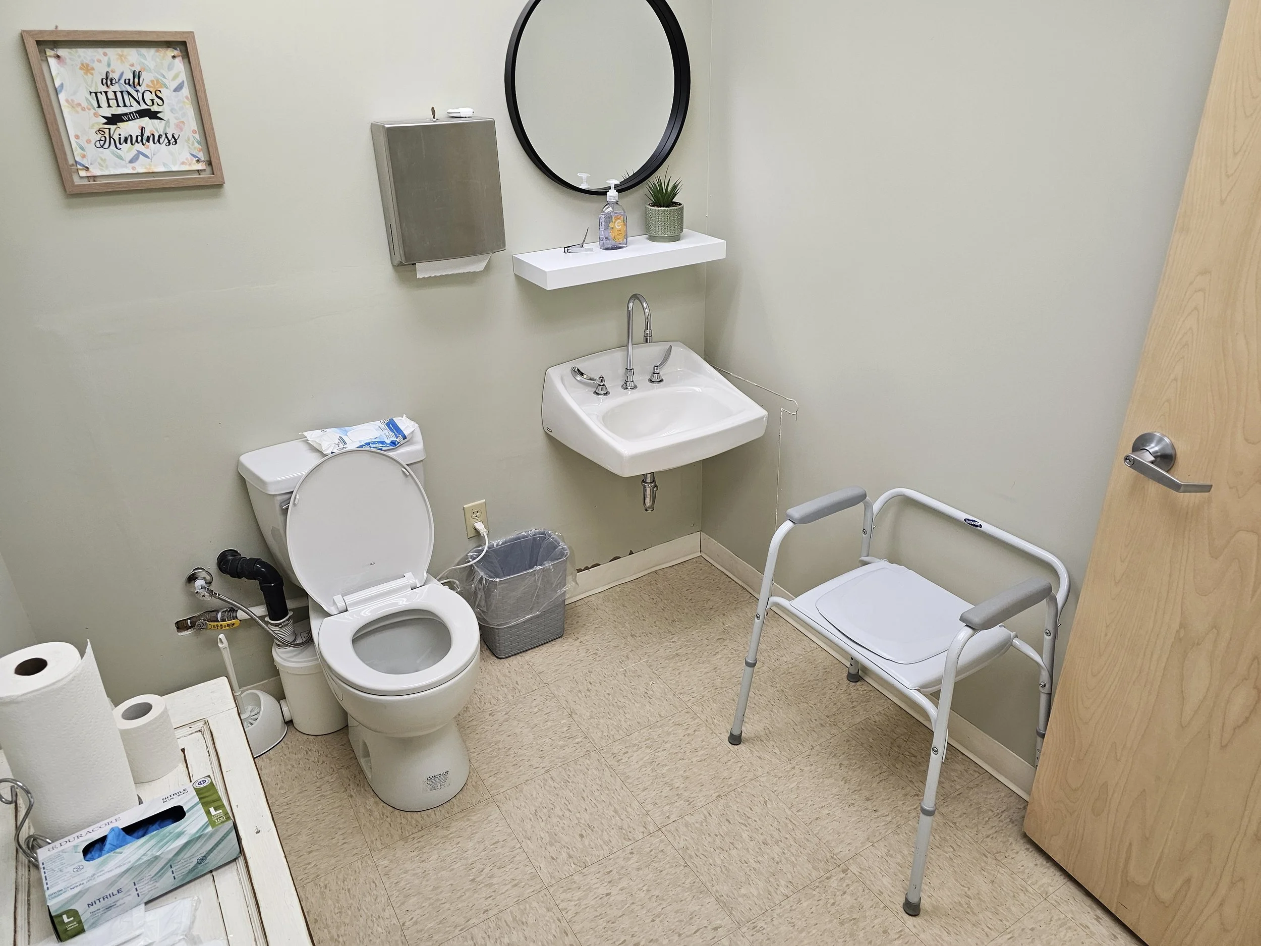 A clean medical office bathroom with a toilet, sink, mirror, wall art, and a walker. Toiletries and paper towels are on a small white table. There's a trash bin and hand sanitizer on a shelf.