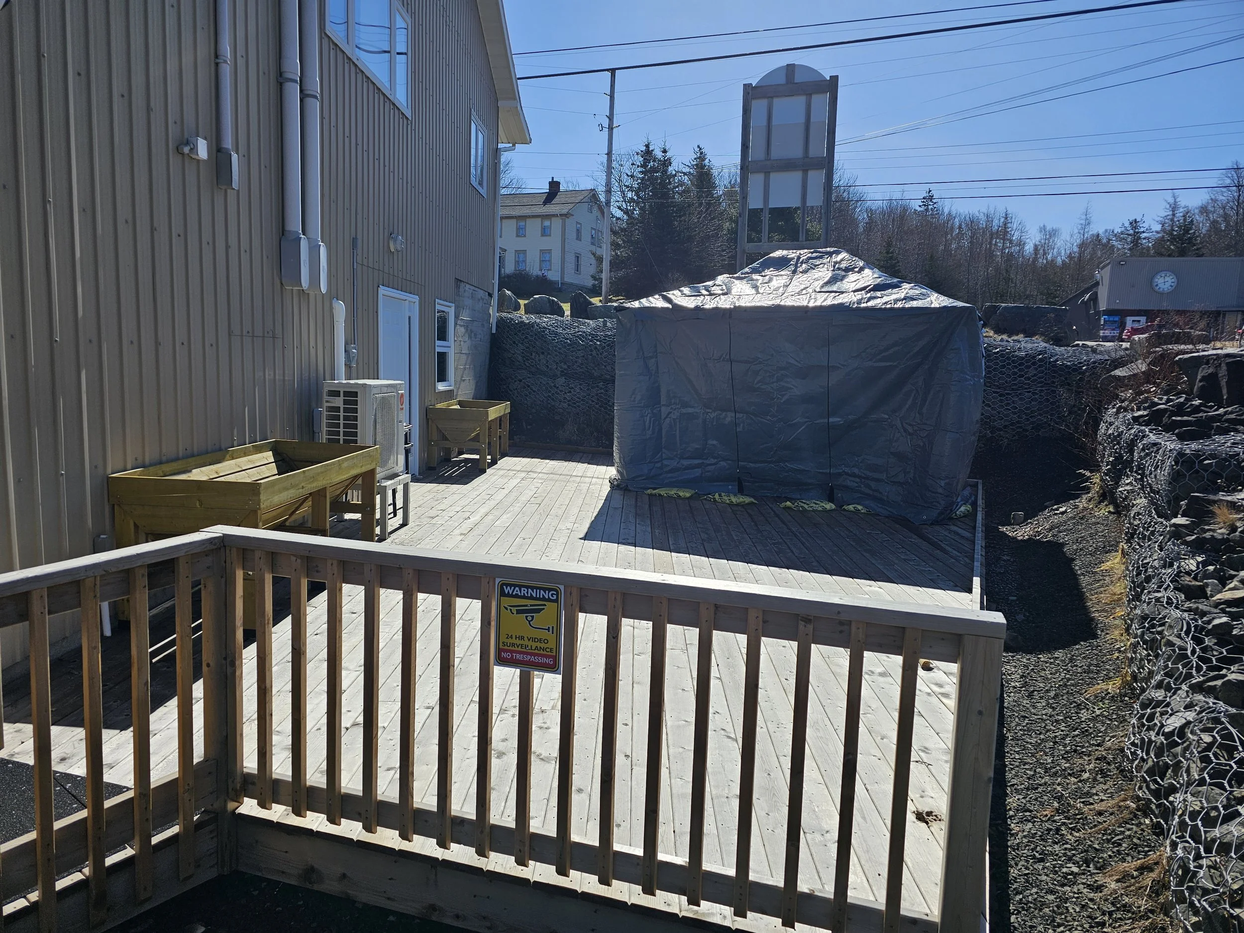 A wooden deck attached to a building with two planter boxes, an air conditioning unit, and a covered object. There is a warning sign on the deck railing about video surveillance. The background shows houses, trees, and a clear sky.