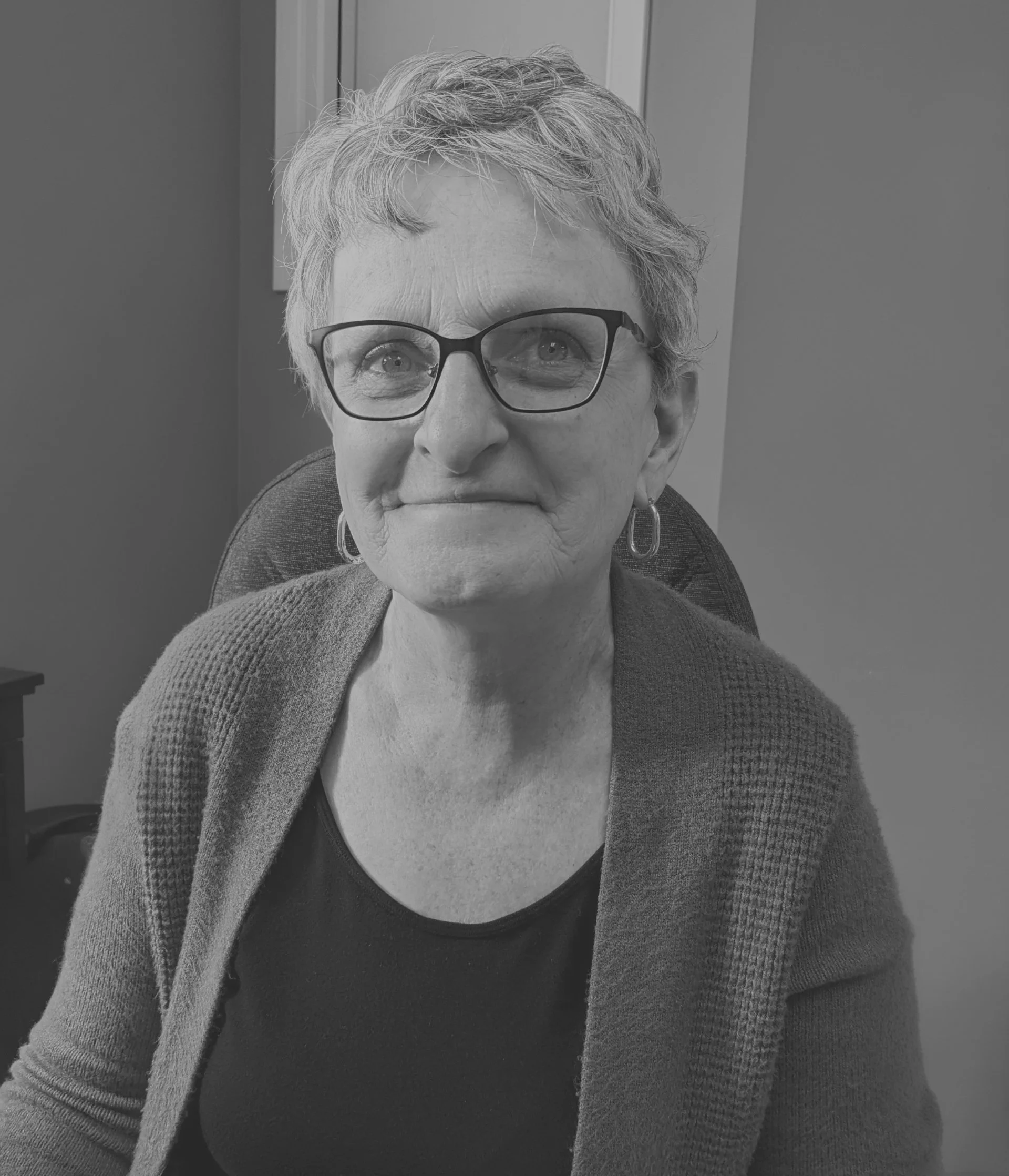 A woman with short, wavy hair, glasses, wearing earrings, a dark top, and a sweater, sitting in an office chair, smiling slightly.