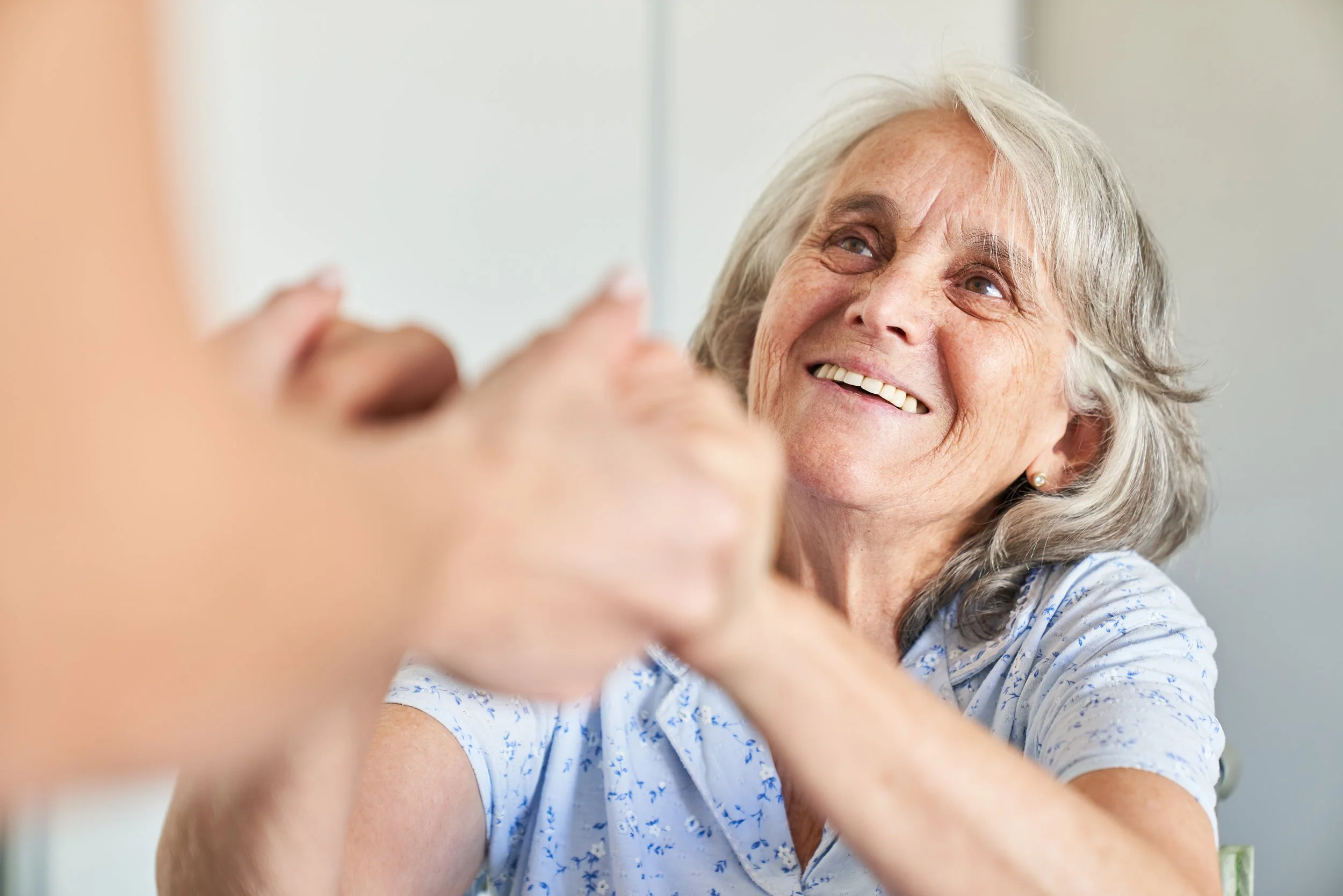 An elderly woman with gray hair smiling and holding hands with another person.