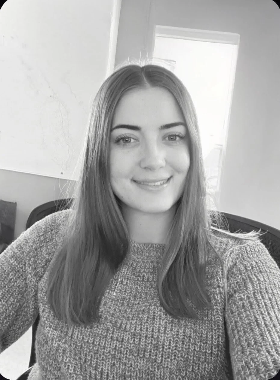 Black and white photo of a young woman with long hair, smiling, sitting in an indoor setting, wearing a knitted sweater.