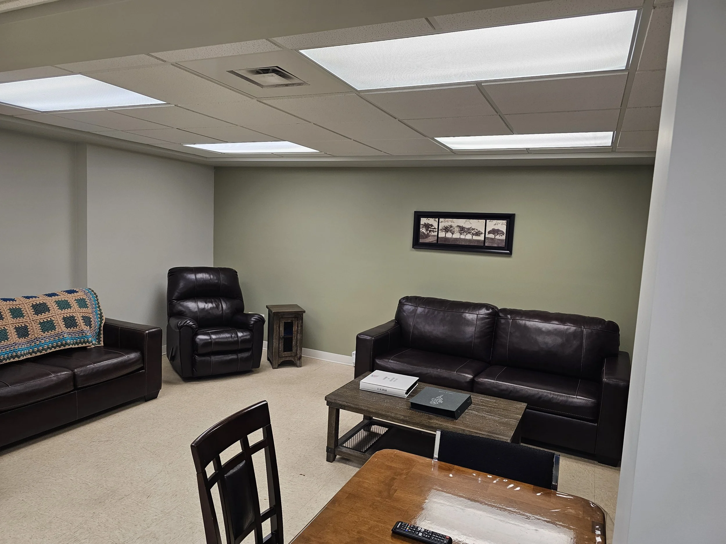 A small, cozy lounge area with two leather sofas, a leather armchair, a coffee table with books, a small side table, a framed picture on the wall, and part of a wooden dining table with a remote control on it.