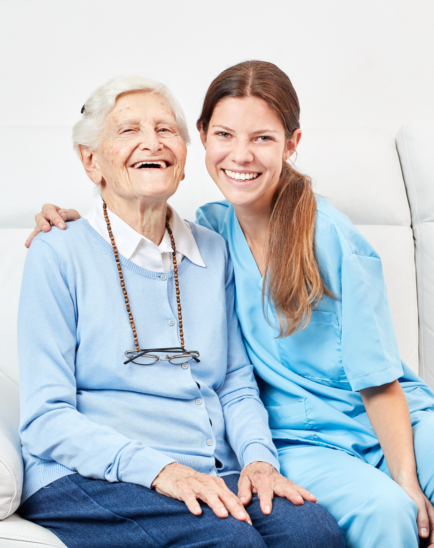 A young woman in medical scrubs sitting beside an elderly woman with a white shirt, cardigan, and glasses hanging around her neck, both smiling and sitting on a white couch.