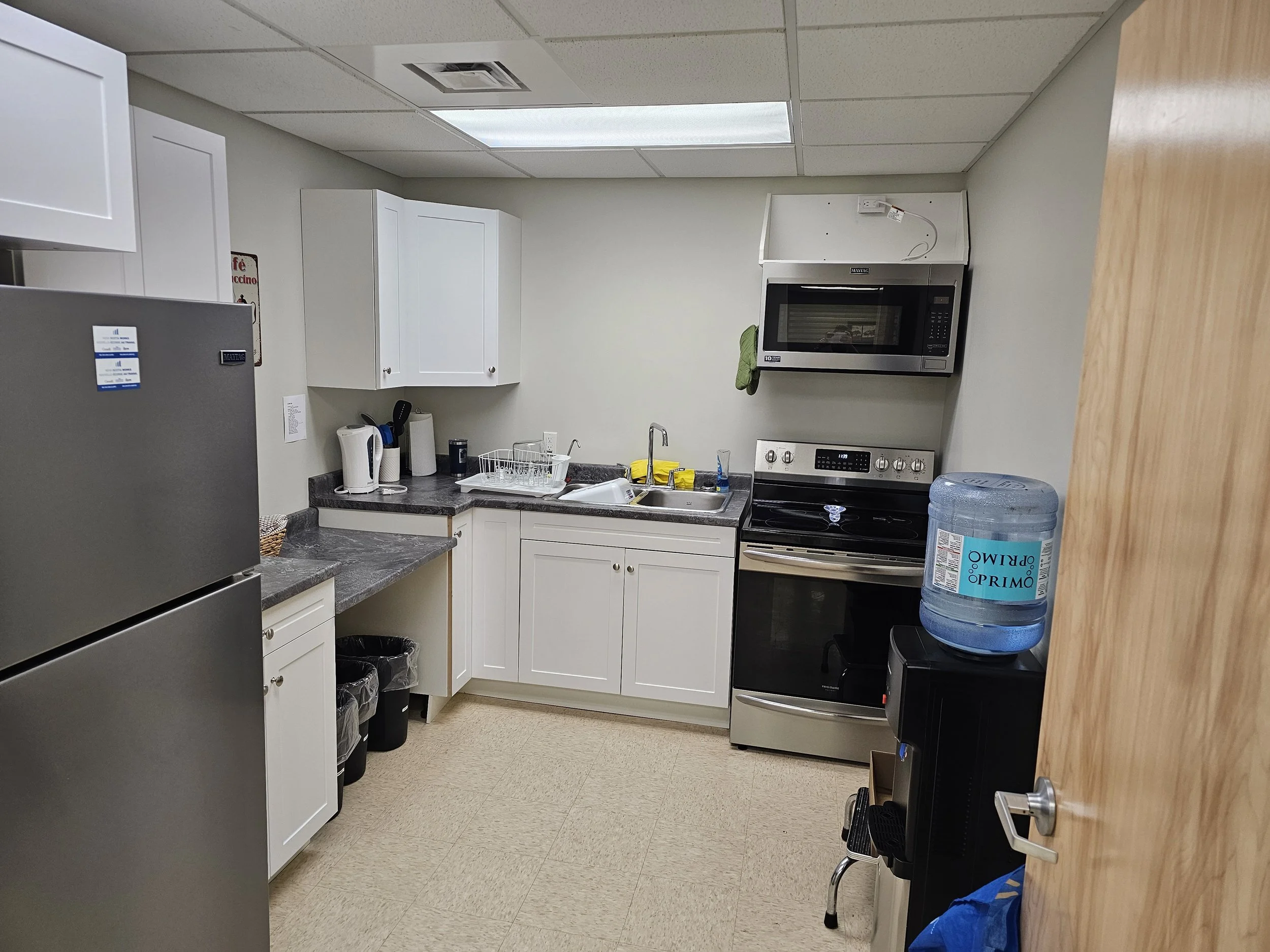 A small kitchen with white cabinets, gray countertops, a stainless steel refrigerator, a microwave above a stove, a water dispenser, and various kitchen appliances and supplies.