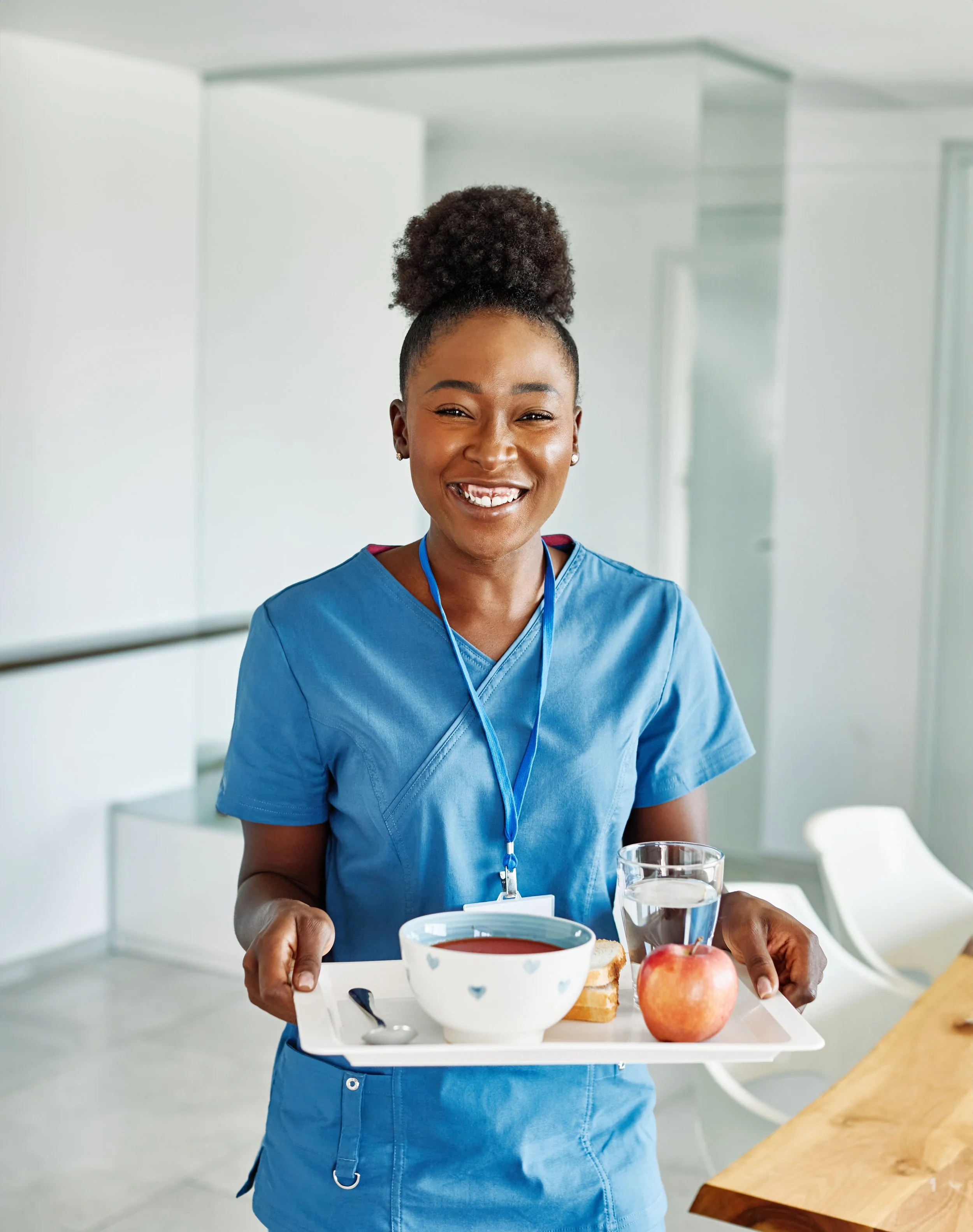 A smiling female healthcare worker in blue scrubs holding a tray with a bowl of soup, a glass of water, an apple, and a slice of bread in a hospital or clinic setting.