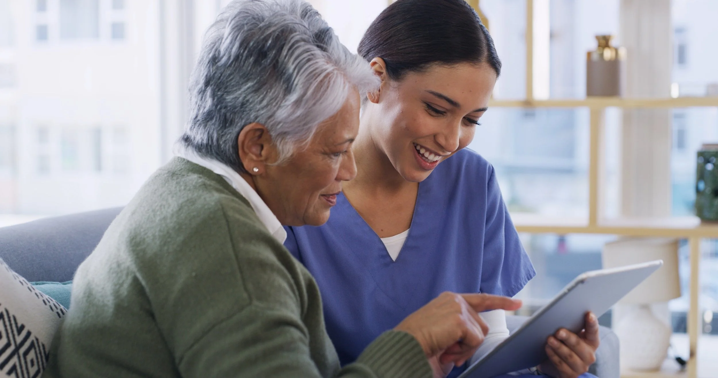 A young female healthcare worker in blue scrubs showing a tablet to an elderly woman with gray hair and a green sweater. They are sitting together on a couch, smiling and looking at the tablet in a bright room with large windows.