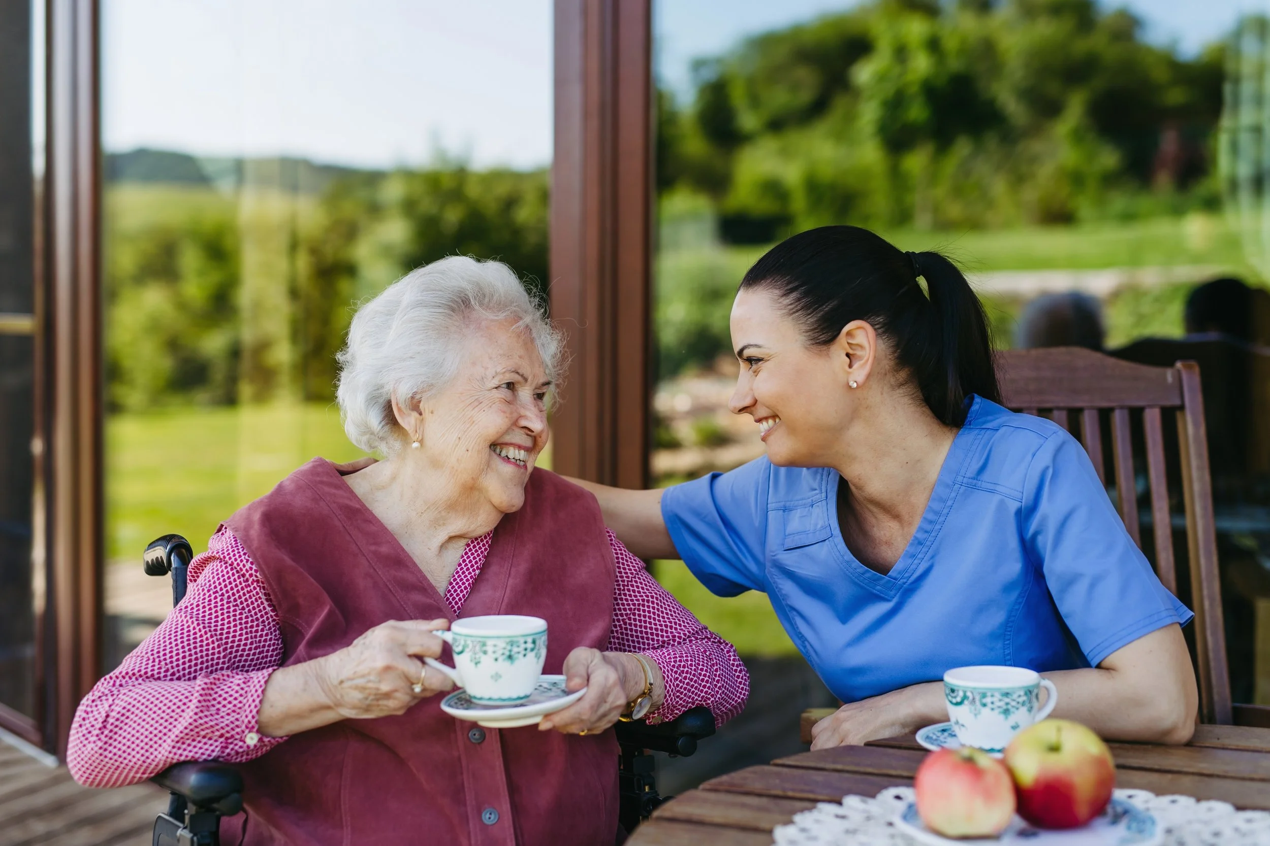 A young nurse smiling at an elderly woman in a wheelchair, holding a teacup, while sitting at a wooden table outside with apples and other teacups.