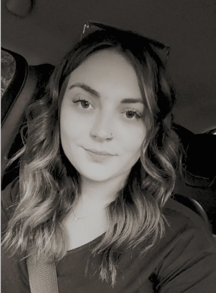 A young woman with wavy hair, wearing sunglasses on her head and a nose piercing, taking a selfie in a car, in black and white.