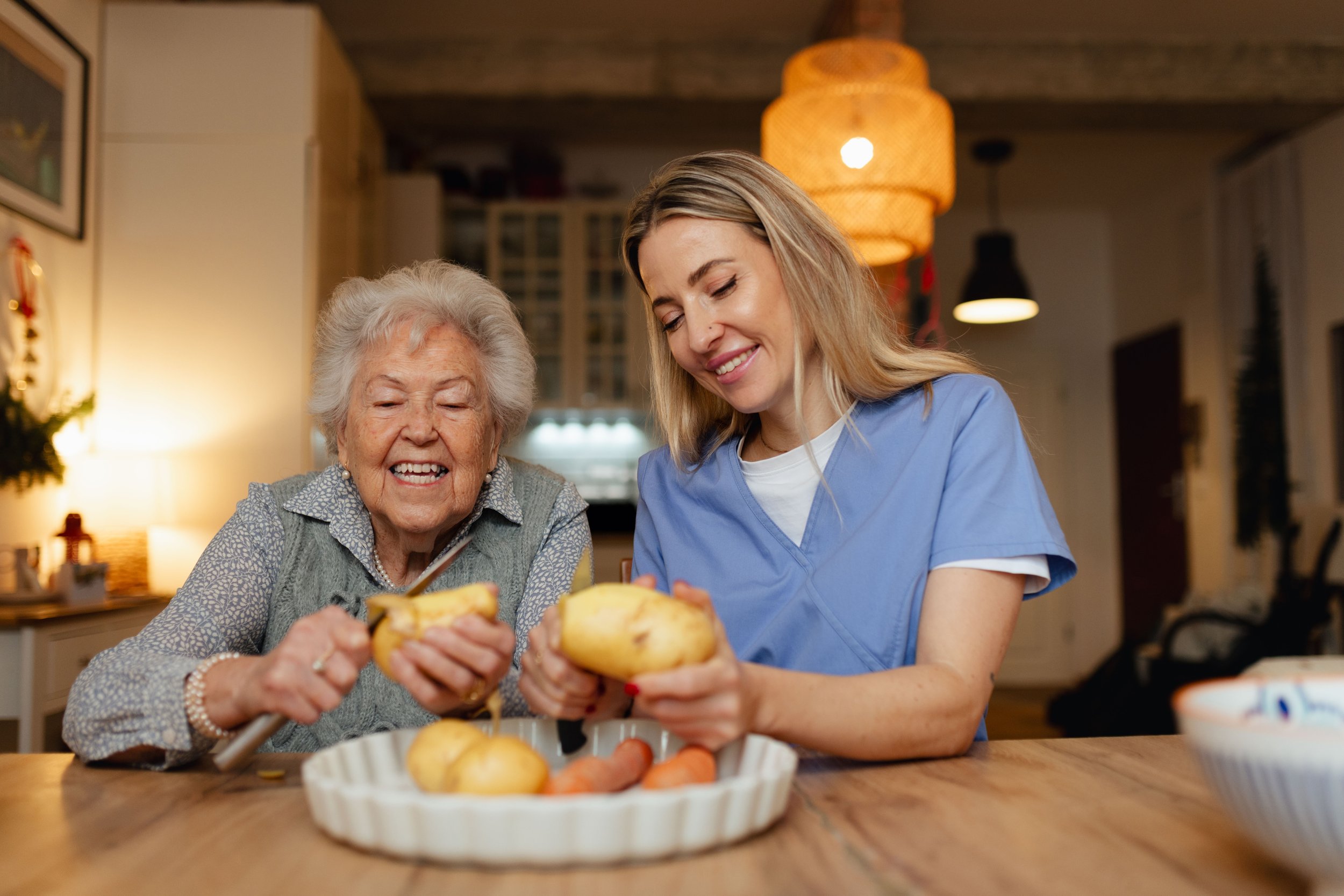 A young caregiver and an elderly woman peeling potatoes together at a kitchen table, smiling and enjoying their time.