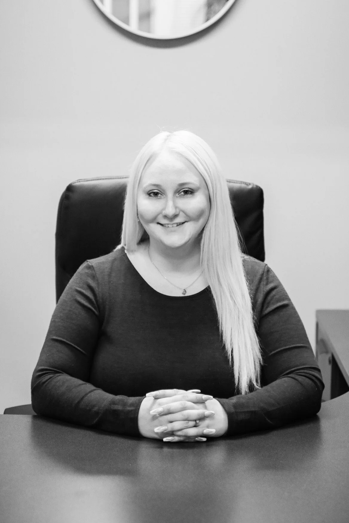 A woman with long blonde hair sitting at a desk, smiling at the camera, in an office setting.