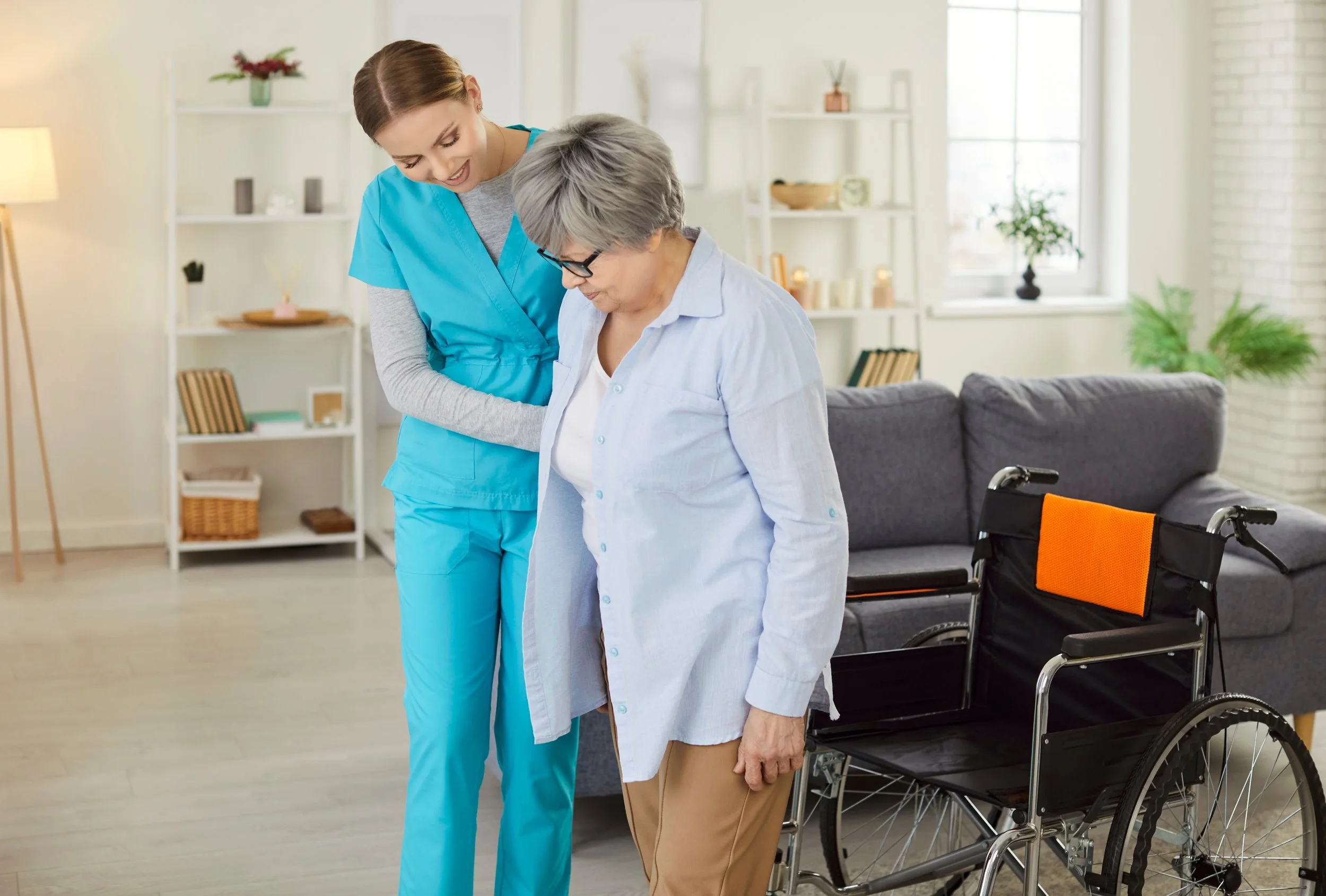 A young female nurse helping an elderly woman with a walking stick in a living room, with a wheelchair nearby.