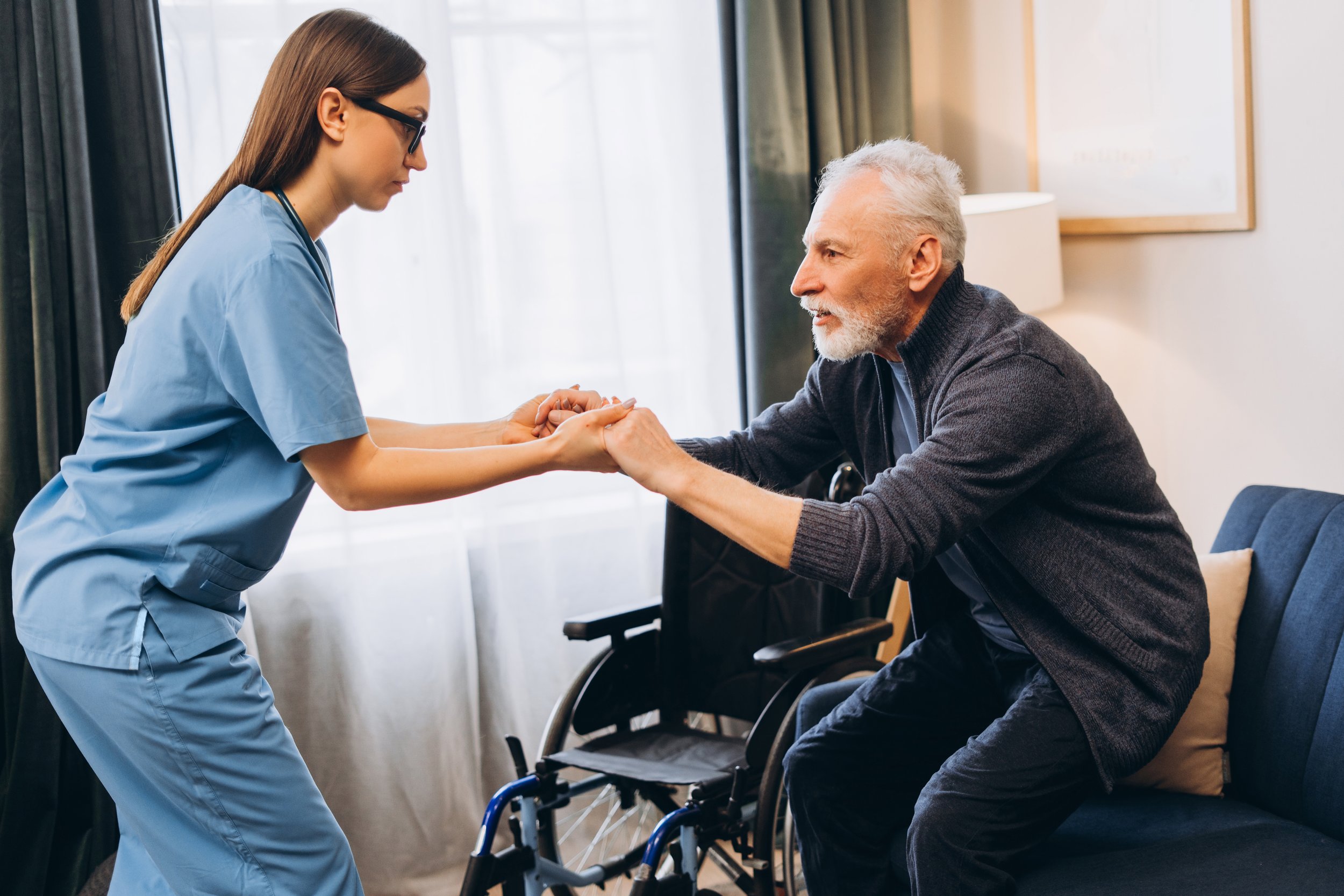 Nurse helping a senior man in a wheelchair during therapy session, holding his hands in a supportive manner.
