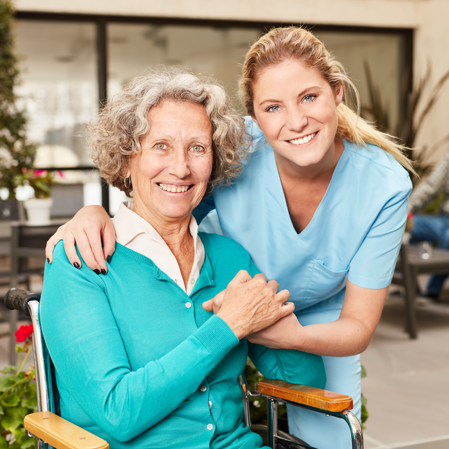 A senior woman in a wheelchair holding hands with a young female caregiver dressed in medical scrubs, both smiling and looking at the camera in a bright indoor setting.