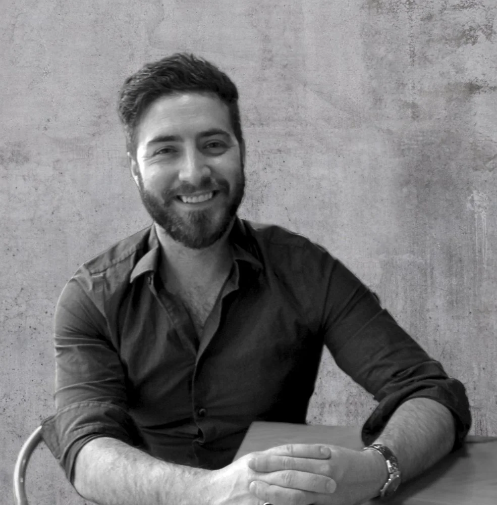 A smiling man with a beard and short hair, wearing a dark button-up shirt, sitting at a table against a textured wall.