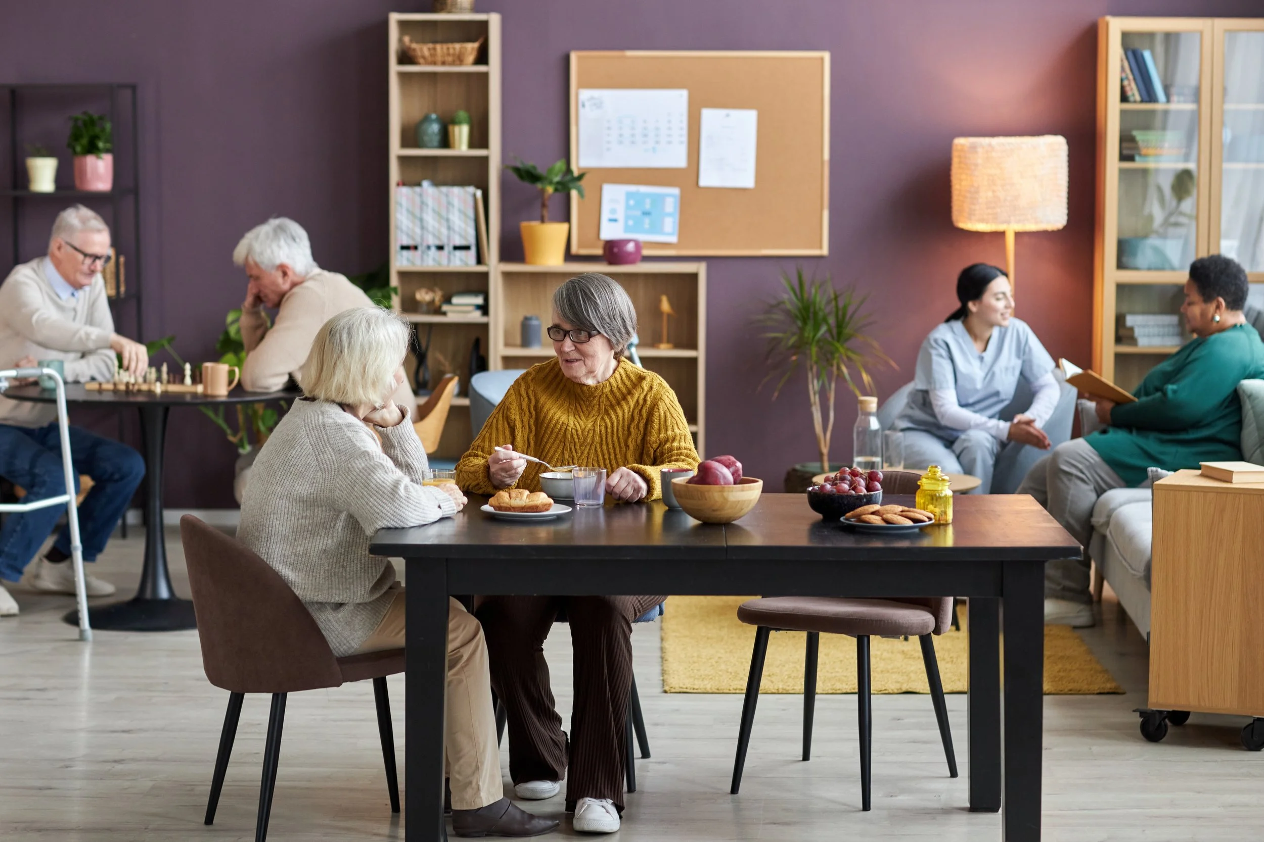 People socializing in a cozy room with wooden shelves, a purple wall, and a dining table with fruit and snacks. Some are talking, others playing chess or reading.