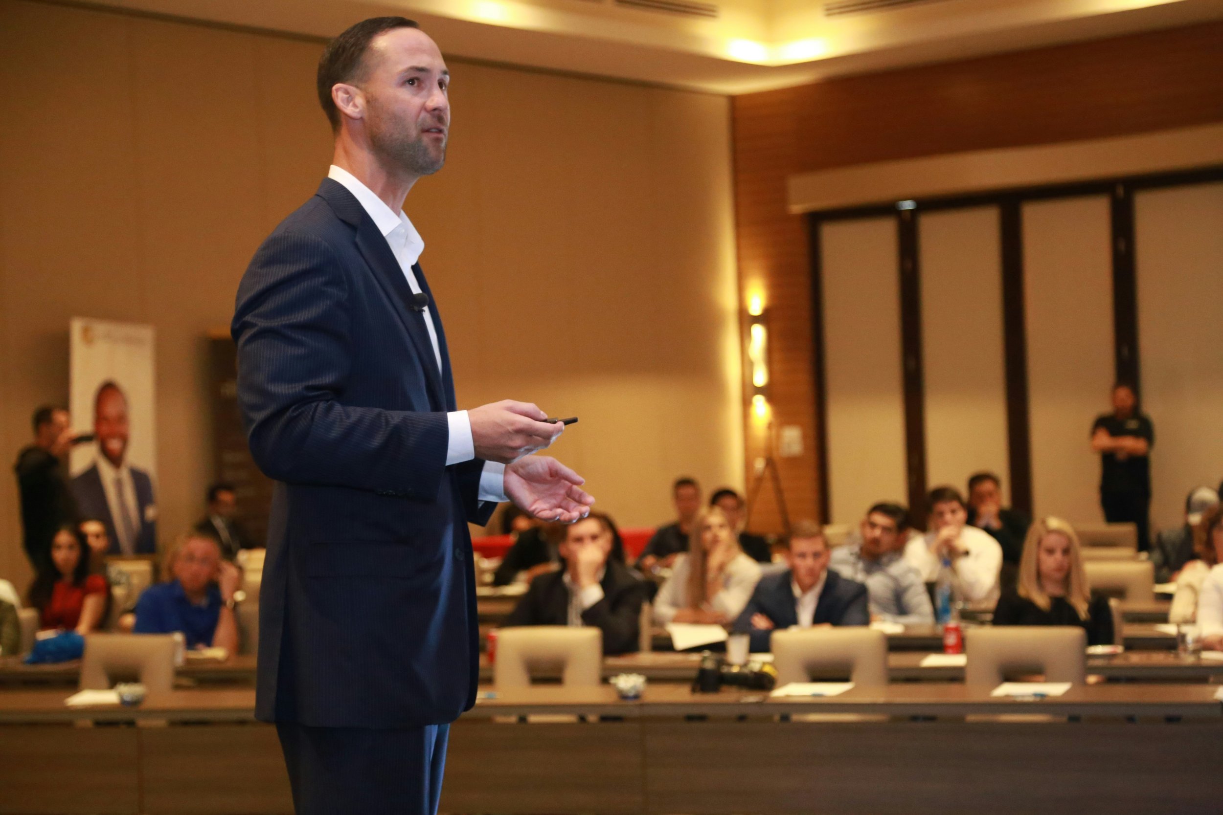 A man in a navy suit giving a presentation to a seated audience in a conference room.