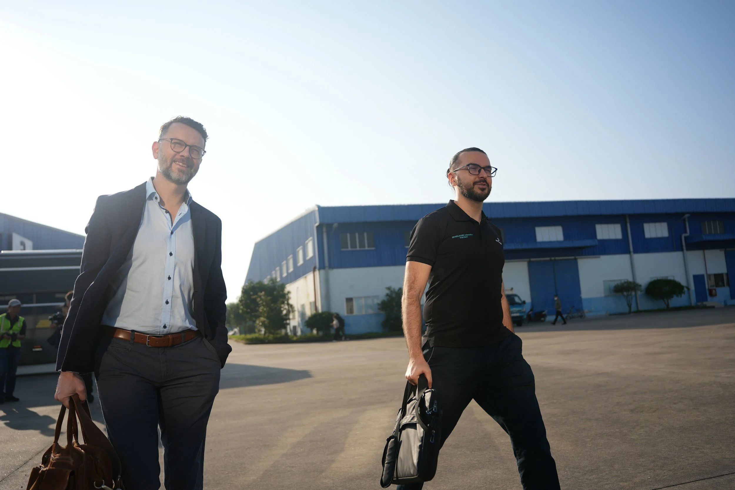 Two men walking outside near a large blue industrial building on a sunny day. One man is dressed in business attire holding a brown leather bag, the other in a black polo with a backpack.