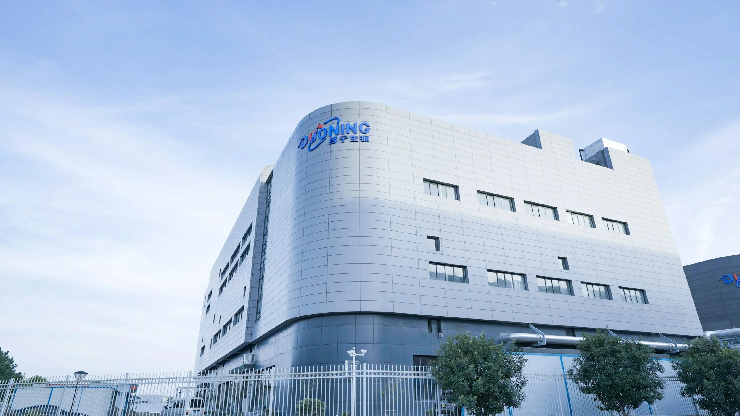 Modern white building with the logo 'DÜNONG' on the top left corner, surrounded by a white fence and green trees, under a partly cloudy sky.