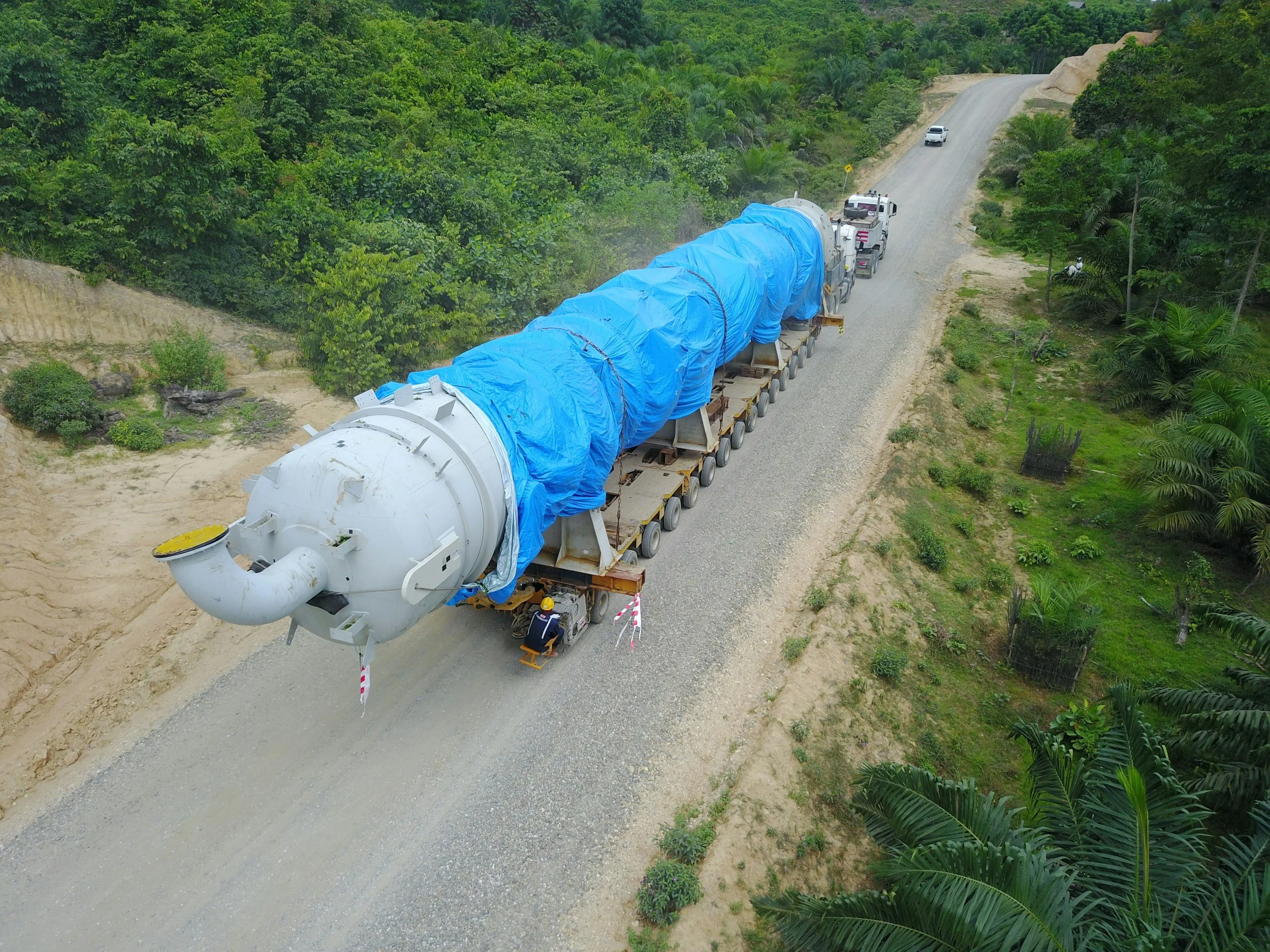 A large industrial equipment or machine wrapped in blue tarp being transported on a flatbed truck along a rural road surrounded by green foliage.