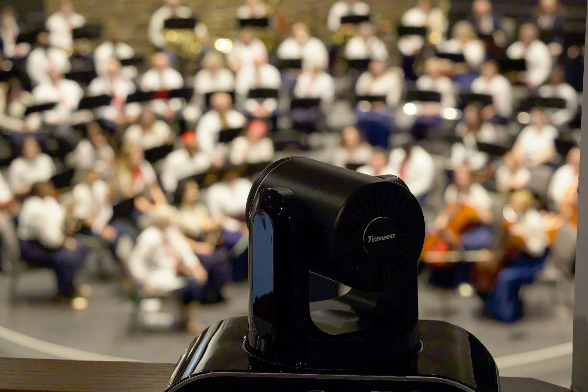 A black speaker with the brand name 'Teneo' on it, placed on a surface in front of a blurred background of an audience, possibly in an auditorium or theater.