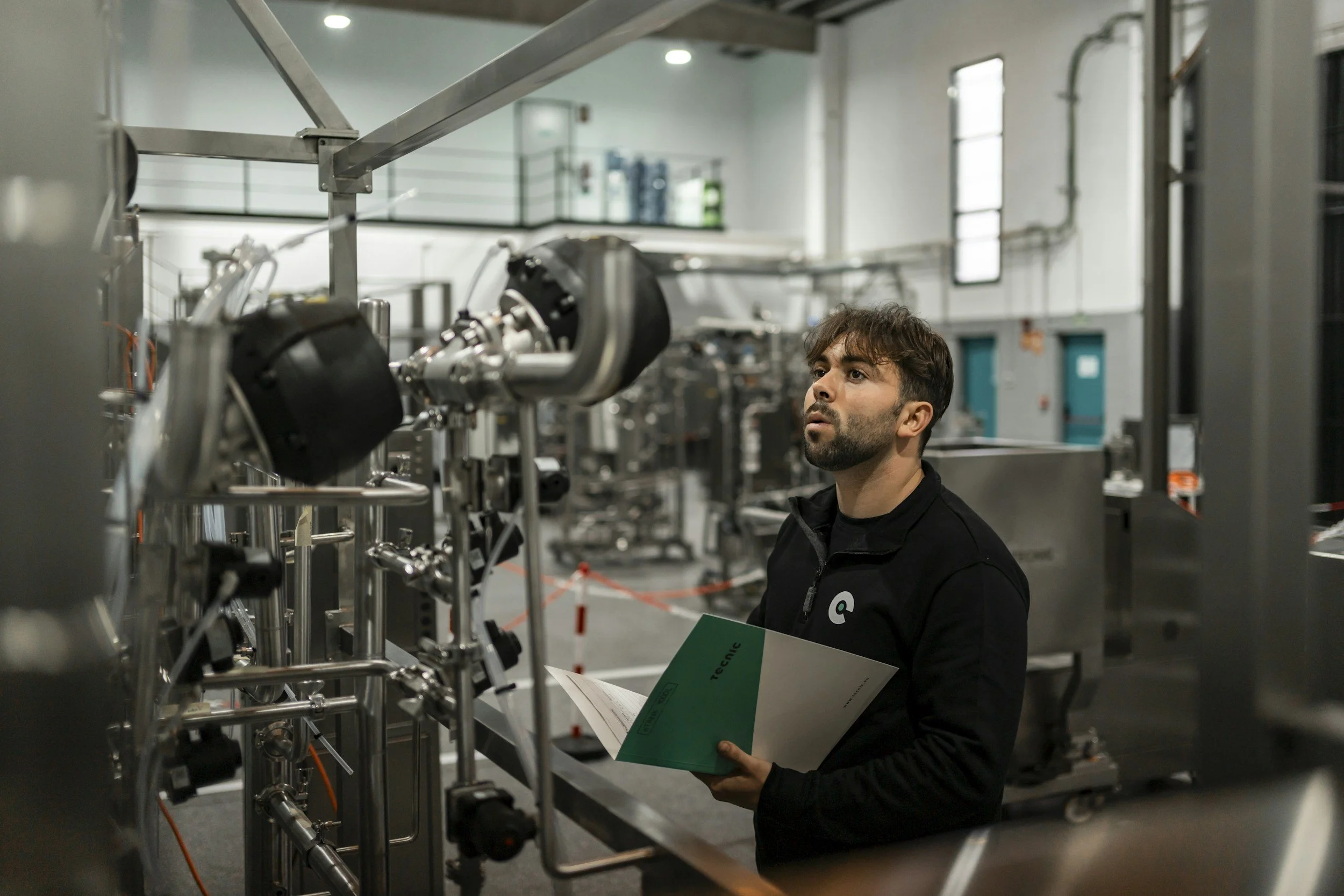 A man with dark hair and beard wearing a black jacket holding a green and white brochure in a laboratory surrounded by stainless steel machinery and equipment.
