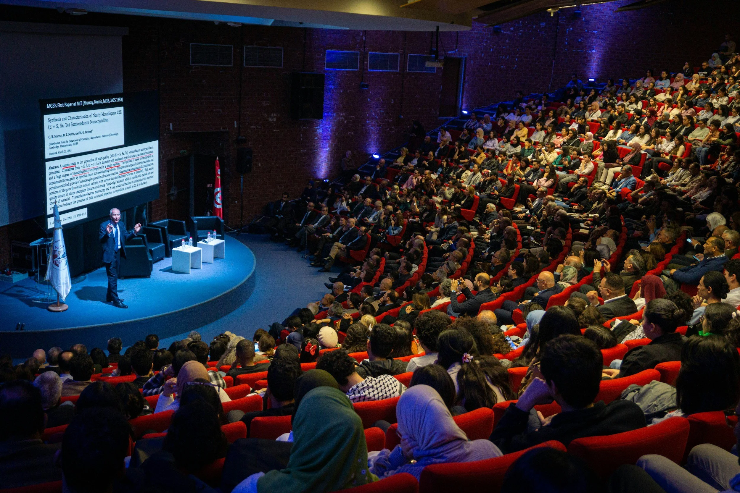 A large audience seated in a theater watching a presentation. A speaker is on stage near a large screen displaying a scientific research slide. The audience consists of diverse individuals, some wearing headscarves, and the setting has red seats and blue and purple lighting on the walls.