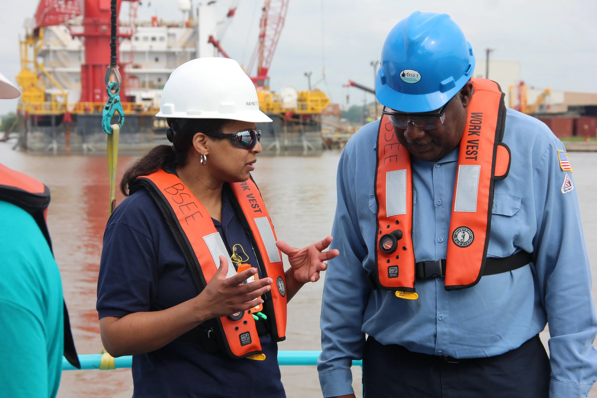 Two workers at a waterfront, wearing hard hats and orange life vests, engaged in a discussion with a large ship and construction equipment in the background.