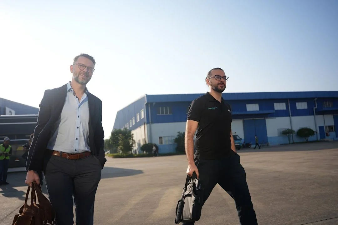 Two men walking outside an industrial building, one dressed in business attire and the other in casual sportswear, carrying bags.