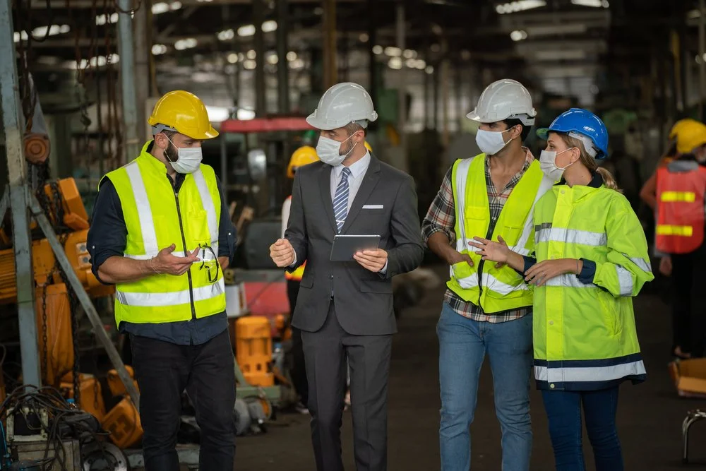 Group of construction workers and a man in a suit having a discussion inside a factory or warehouse, all wearing safety helmets and masks.