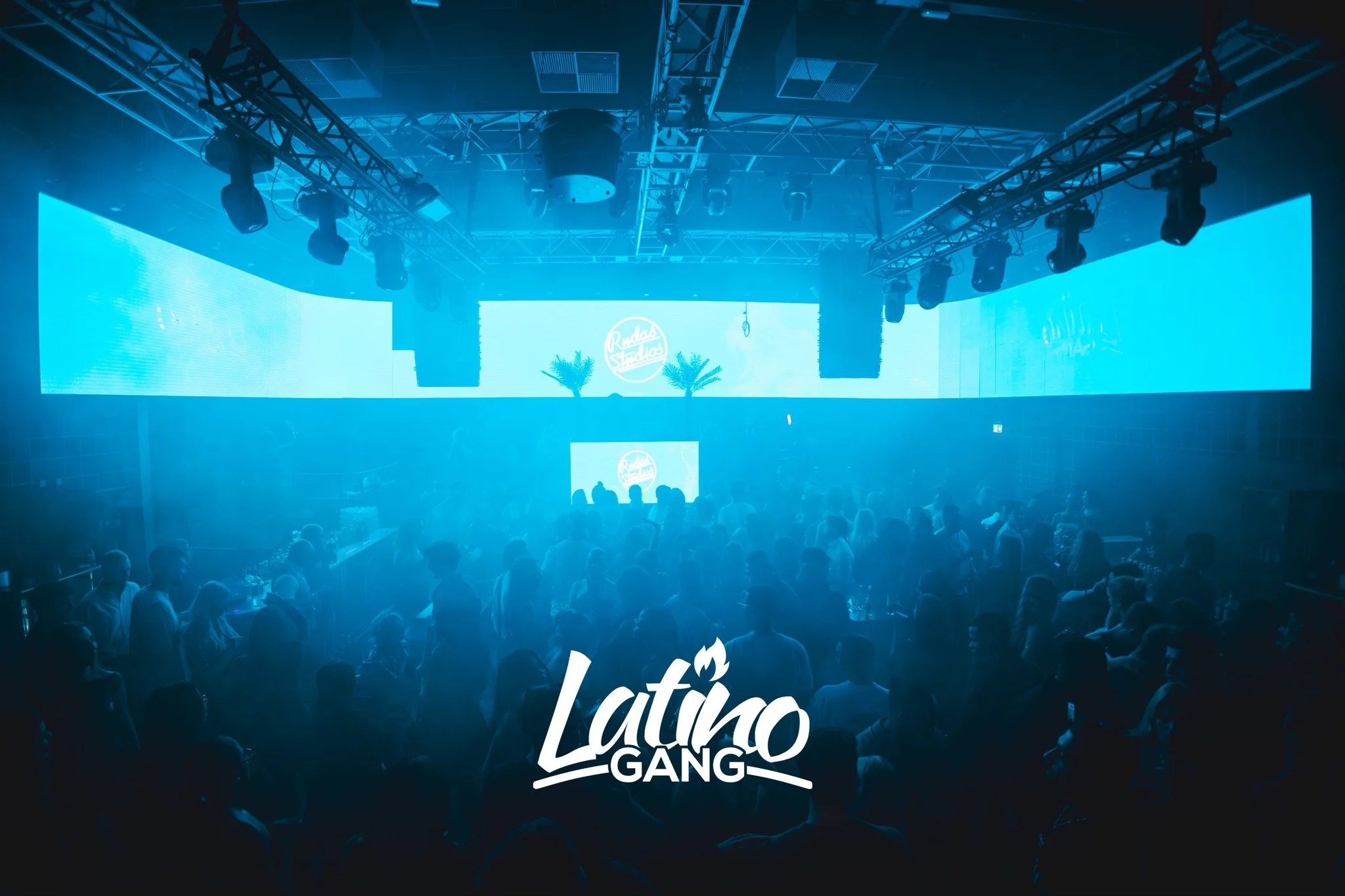 Eine dunkle Clubszene mit blauer Beleuchtung, tanzende Menschen vor einer großen Bühne mit LED-Displays und Palmen, das Logo 'Latino GANG' ist im Vordergrund sichtbar.