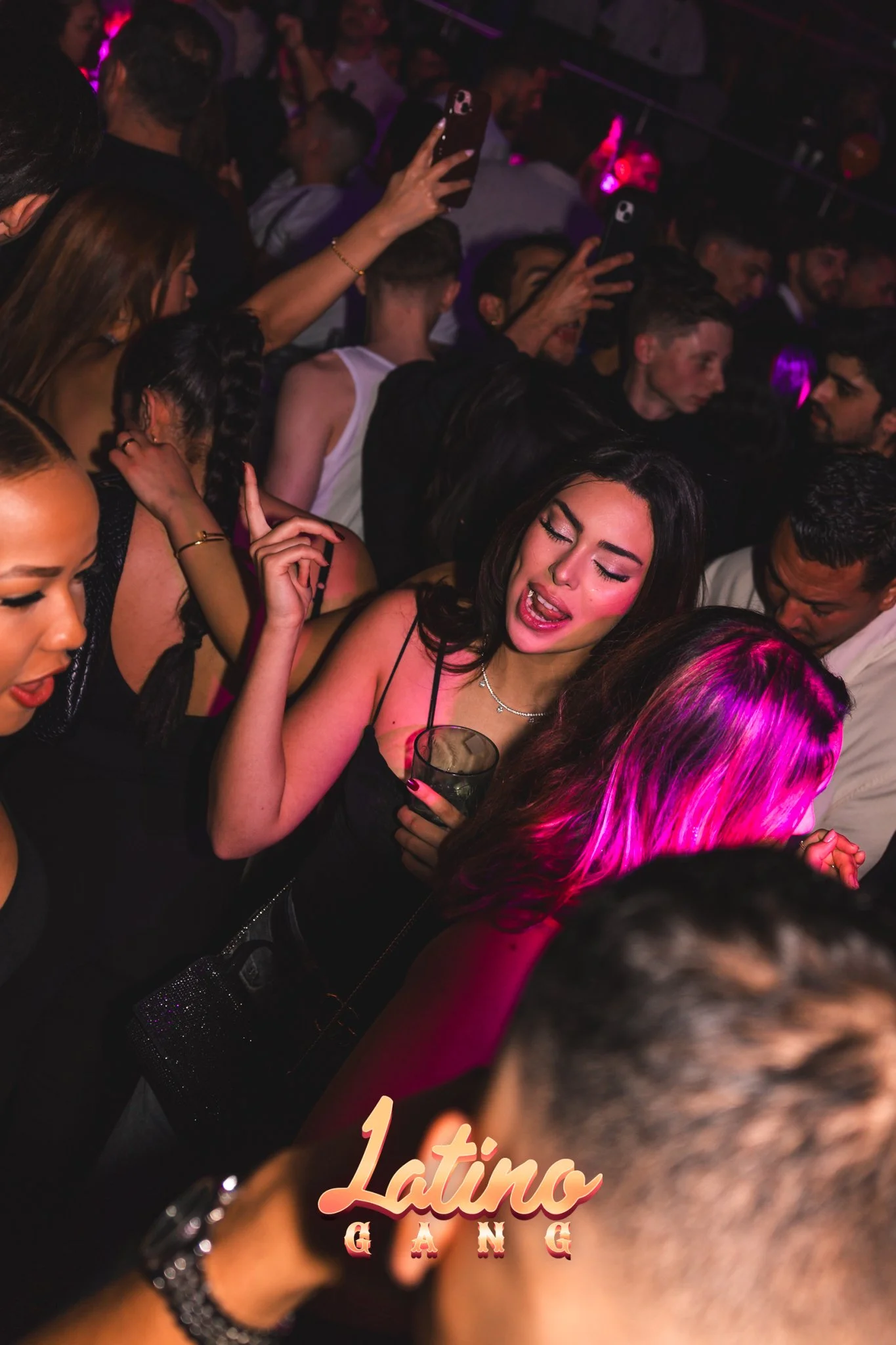 Menschen tanzen und feiern in einer dunklen Club-Umgebung mit bunten Lichtern, hervorstehend eine junge Frau, die singt und eine Hand nach oben hält, während sie ein Getränk hält. Das Bild ist mit dem Text 'Latino GANG' versehen.