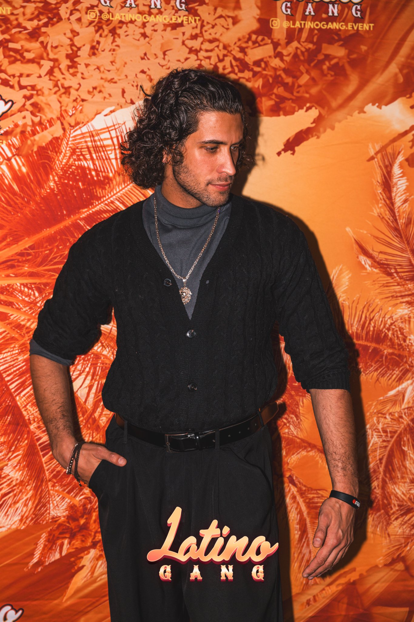 Ein junger Mann mit langen, gewellten dunklen Haaren trägt einen schwarzen Cardigan, einen grauen Rollkragenpullover sowie Schmuck, und steht vor einer orangefarbenen, tropischen Hintergrundwand mit der Aufschrift "Latino GANG" bei einer Veranstaltung.