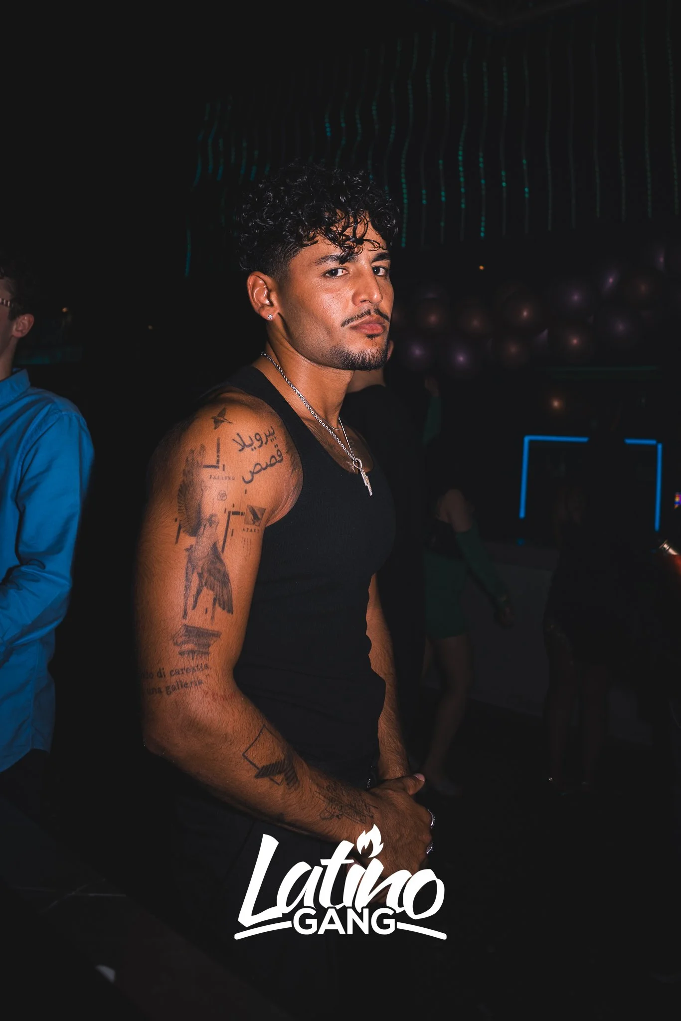 Ein Mann mit dunklen Locken und Tattoos auf dem rechten Arm, trägt ein schwarzes Tanktop in einem dunklen Club, mit dem Text "Latino GANG" im Vordergrund.