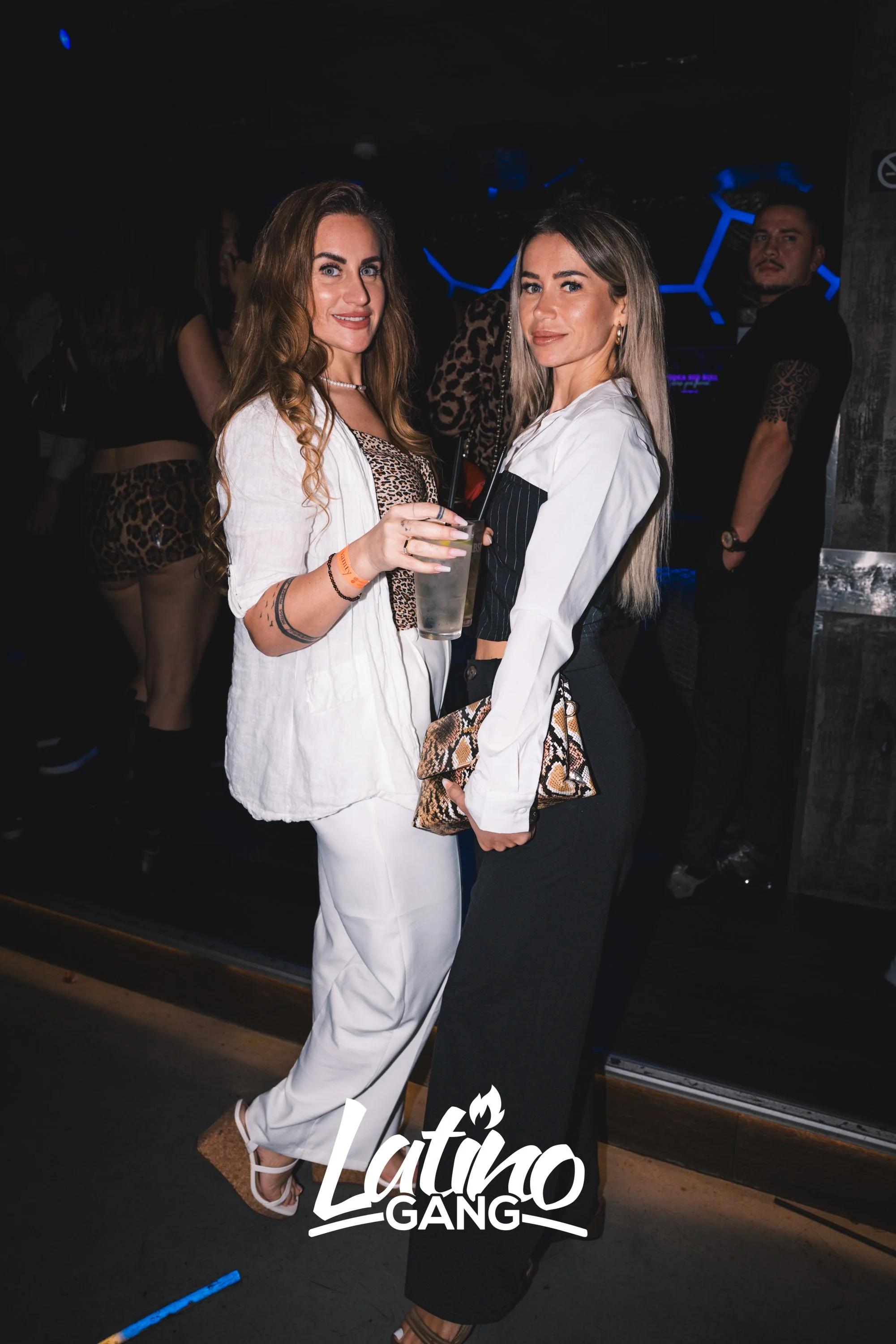 Zwei Frauen posieren in einem Club, eine hält ein Getränk in der Hand, im Hintergrund sind Menschen mit Tiermuster-Kleidung zu sehen. Das Logo 'Latino GANG' ist am unteren Bildrand sichtbar.