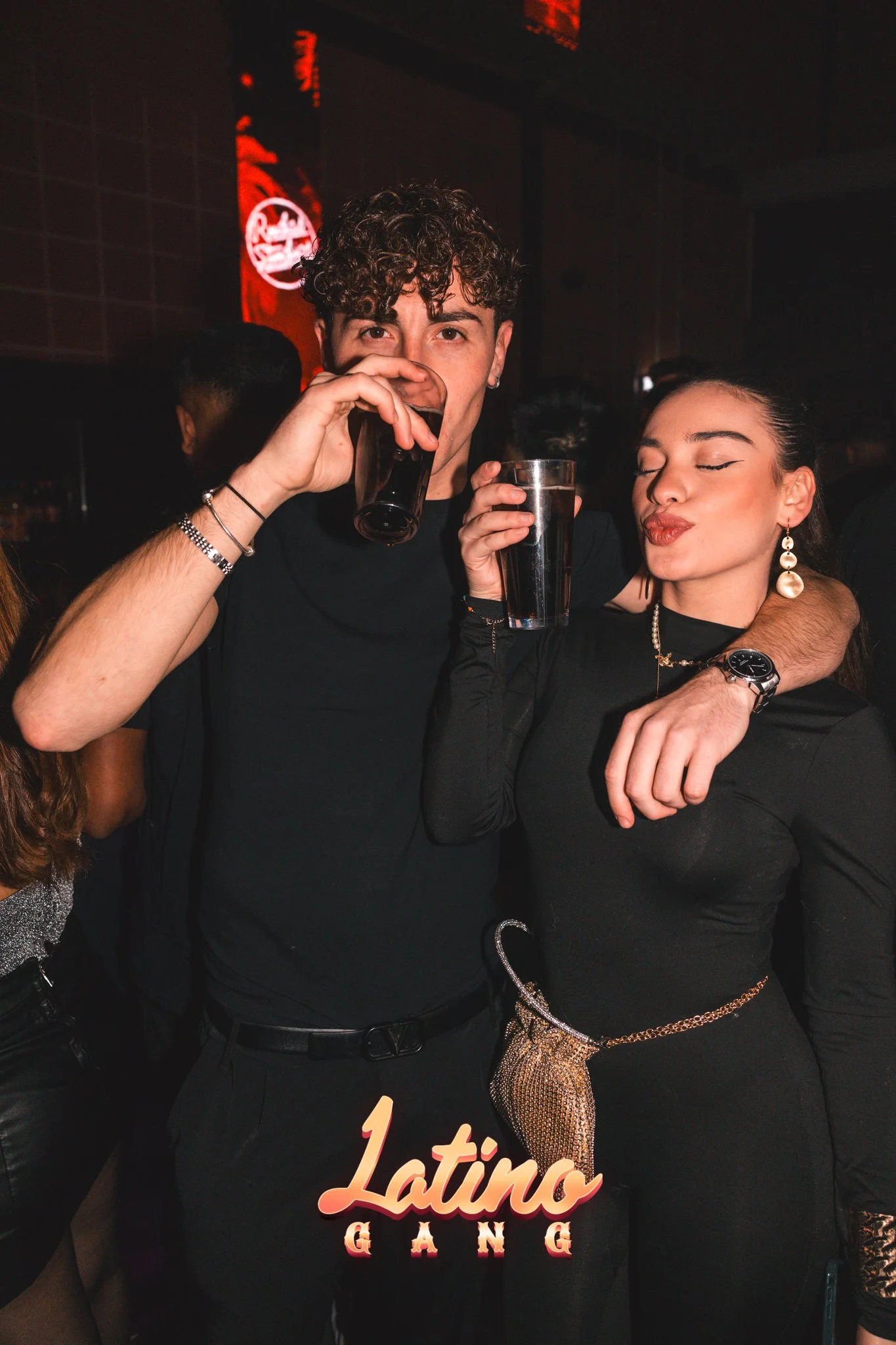 Ein junger Mann und eine junge Frau feiern in einem dunklen Raum, trinken Getränke, umarmen sich und machen einen Kussmund. Das Bild hat den Text 'Latino GANG' im Vordergrund.