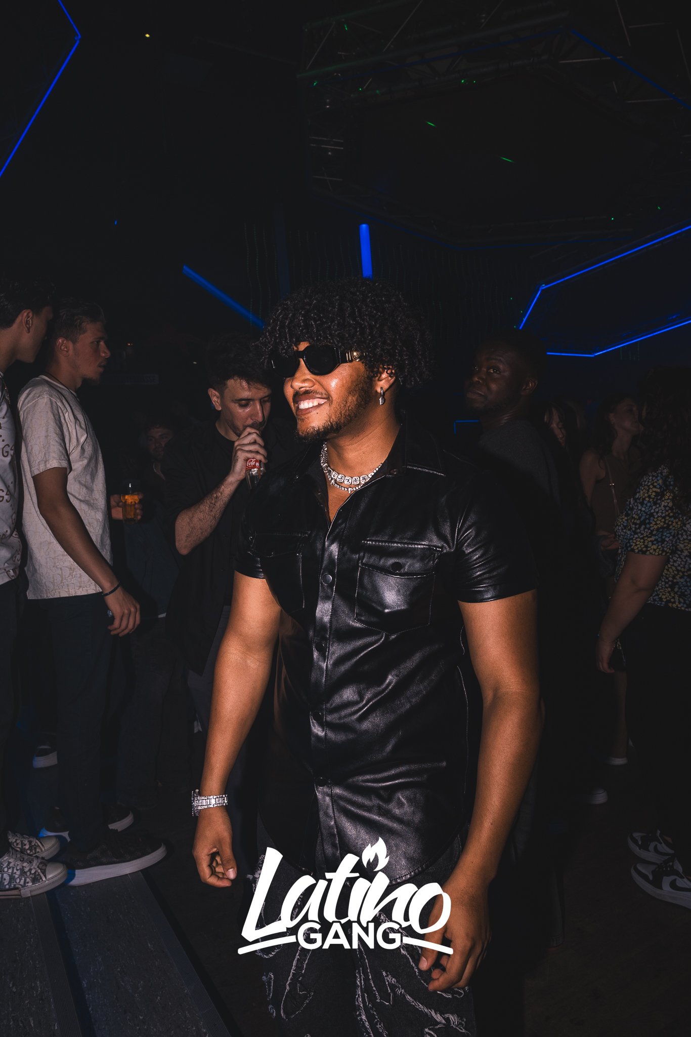 Ein Mann mit lockigem Haar, dunklen Sonnenbrillen und Schmuck trägt ein schwarzes Lederoutfit und tanzt auf einer Party. Im Hintergrund sind weitere Menschen und dunkel leuchtende Neonlichter.