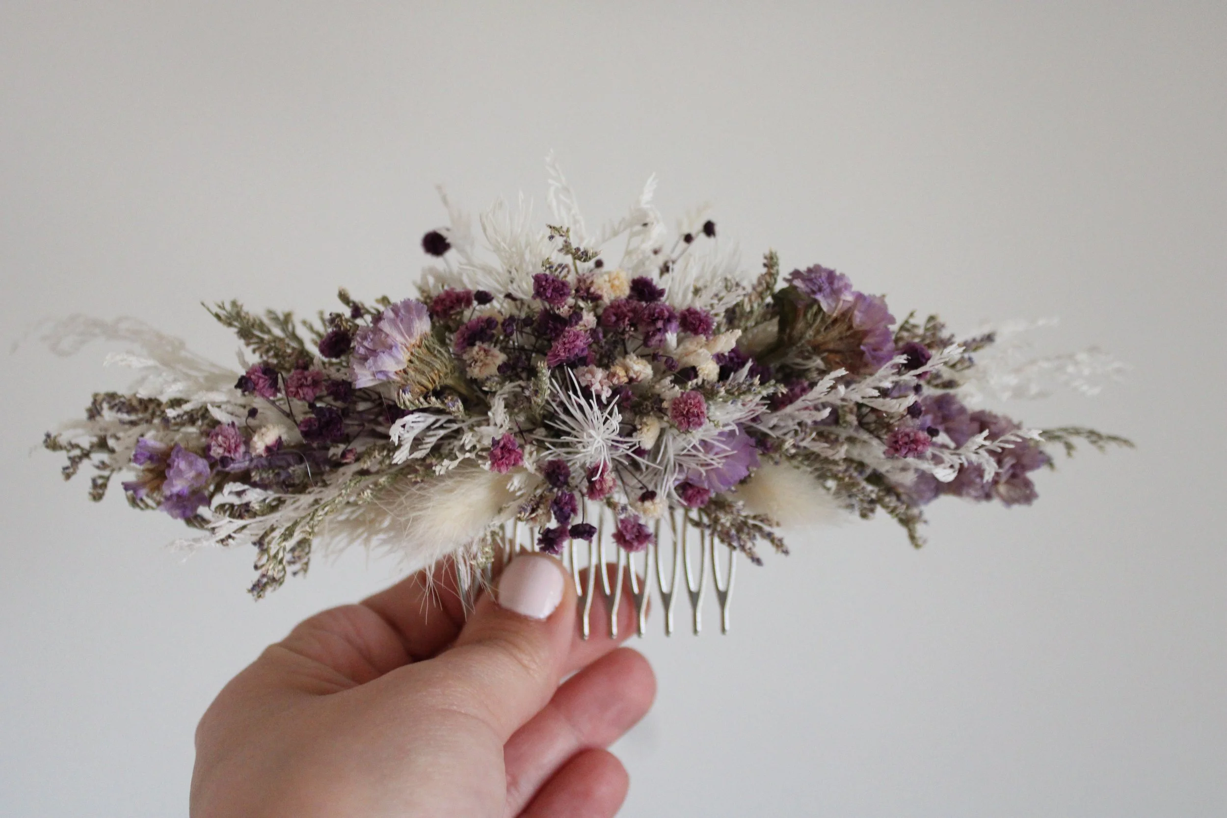 Hand holding a decorative hair comb adorned with dried purple and white flowers and grasses.